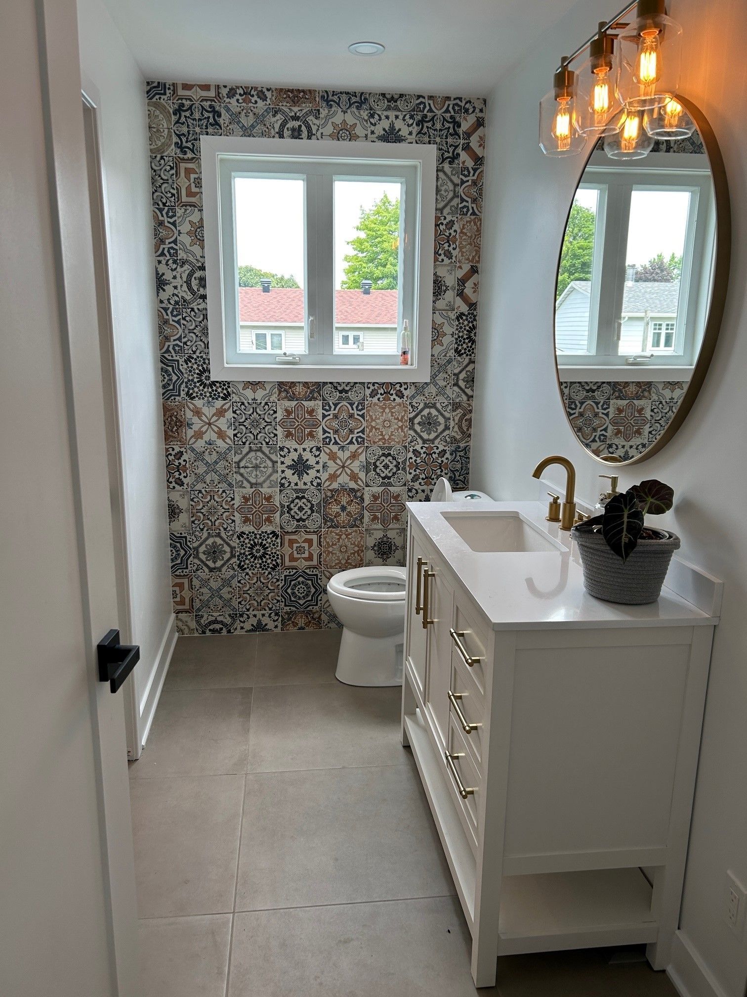 Salle de bain avec mur en carrelage à motifs, vanité blanche, luminaires dorés et fenêtre.