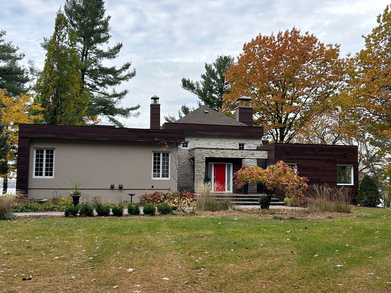 Maison avec murs en pierre et beiges, porte rouge et garniture marron, arbres d'automne en arrière-plan.