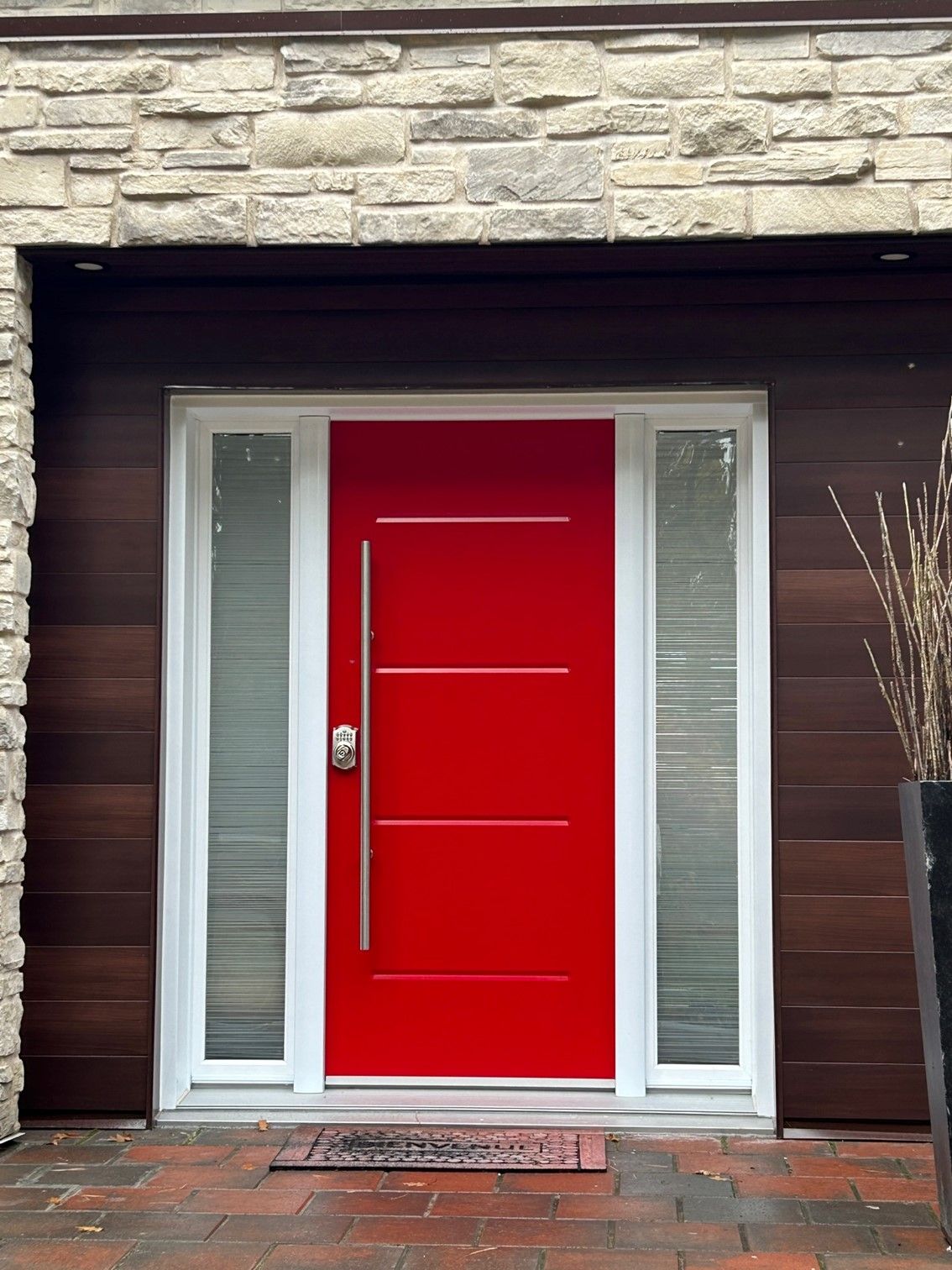 Porte d'entrée rouge avec fenêtres latérales et poignée verticale. Allée en brique, revêtement en pierre et bois.