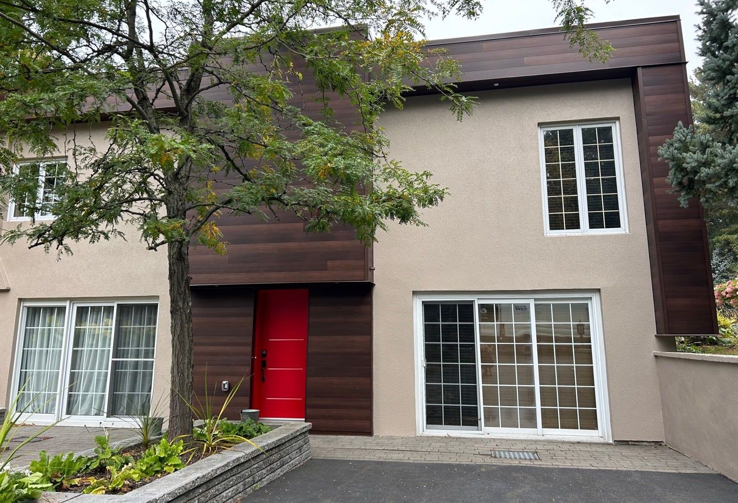 Maison beige à deux étages avec des touches de bois foncé et une porte d'entrée rouge. Un arbre se trouve devant.