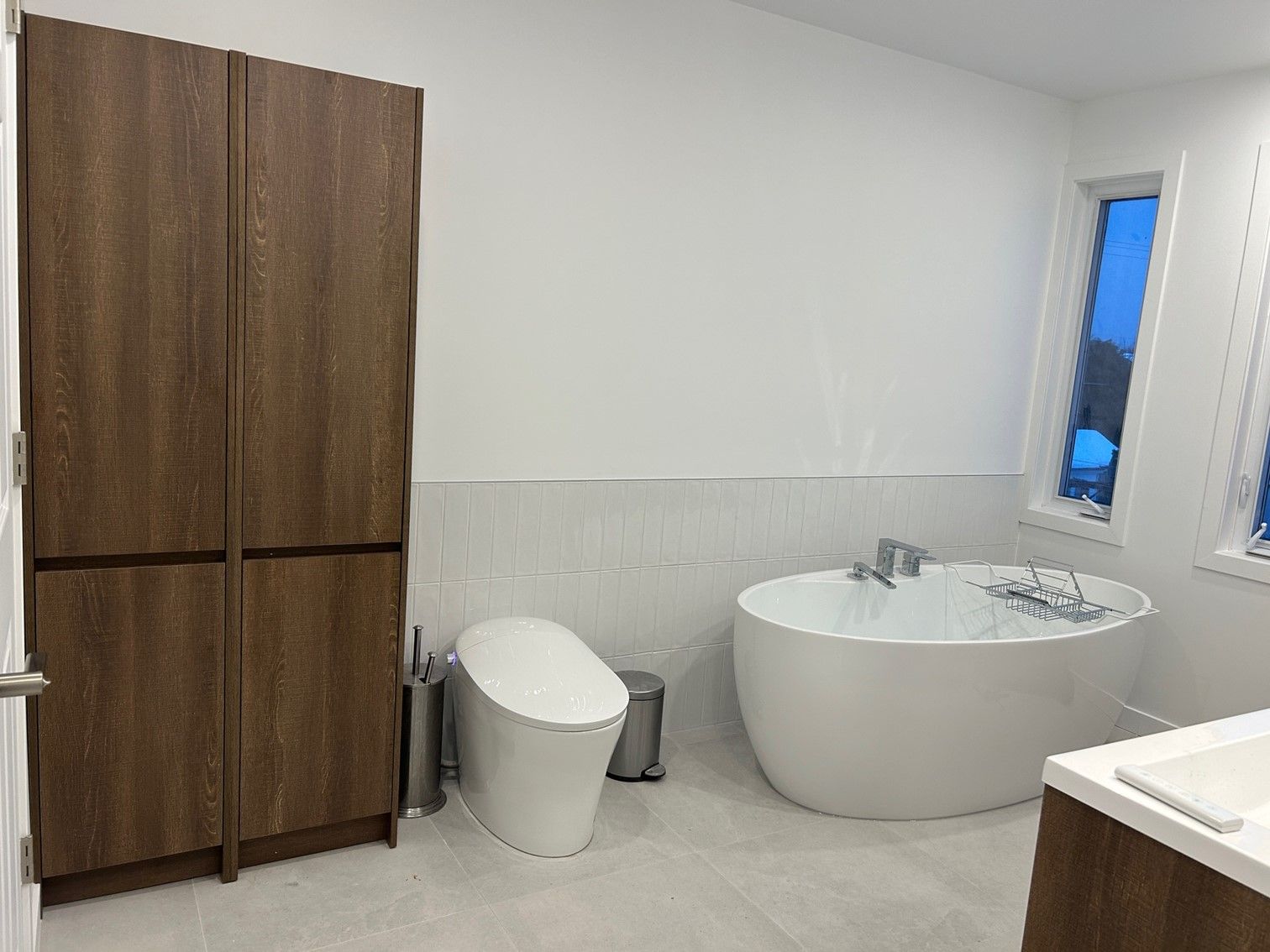 Salle de bain avec baignoire blanche, toilettes et meuble de rangement en bois. Fenêtres laissant entrer la lumière naturelle.