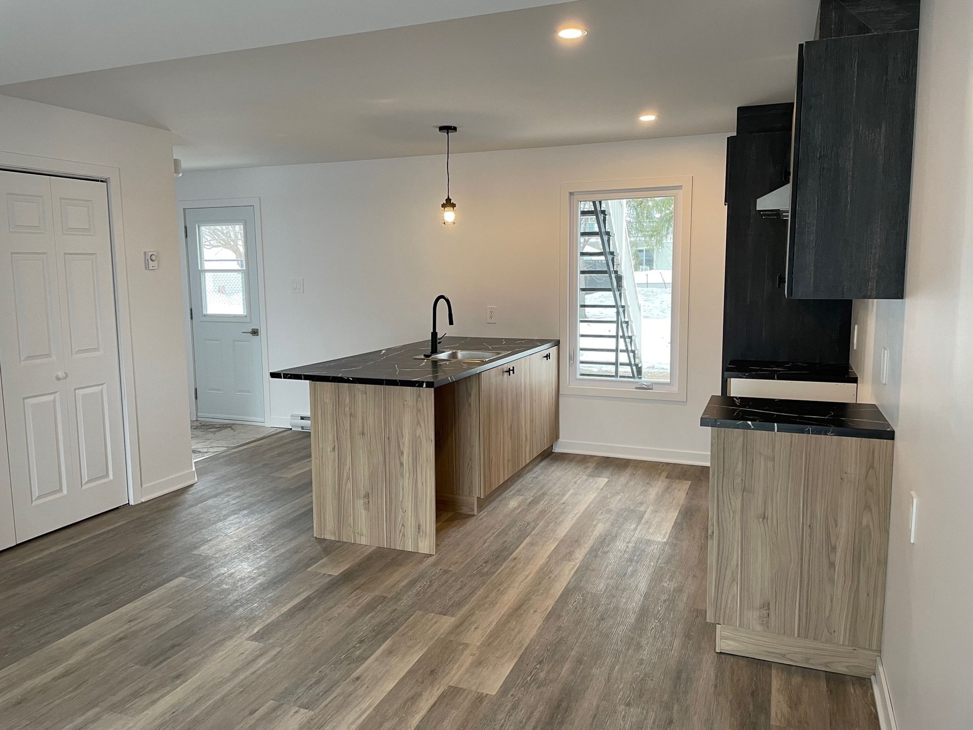 Cuisine moderne avec armoires en bois, comptoirs noirs, îlot avec évier et plancher de couleur claire.