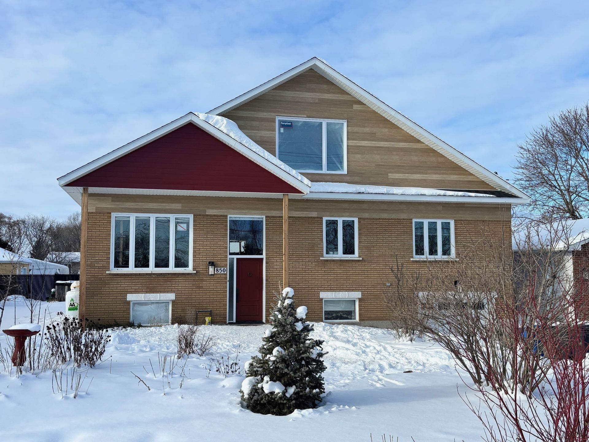 Maison en briques à deux étages avec cour enneigée; auvent rouge, arbre à feuilles persistantes et ciel bleu.