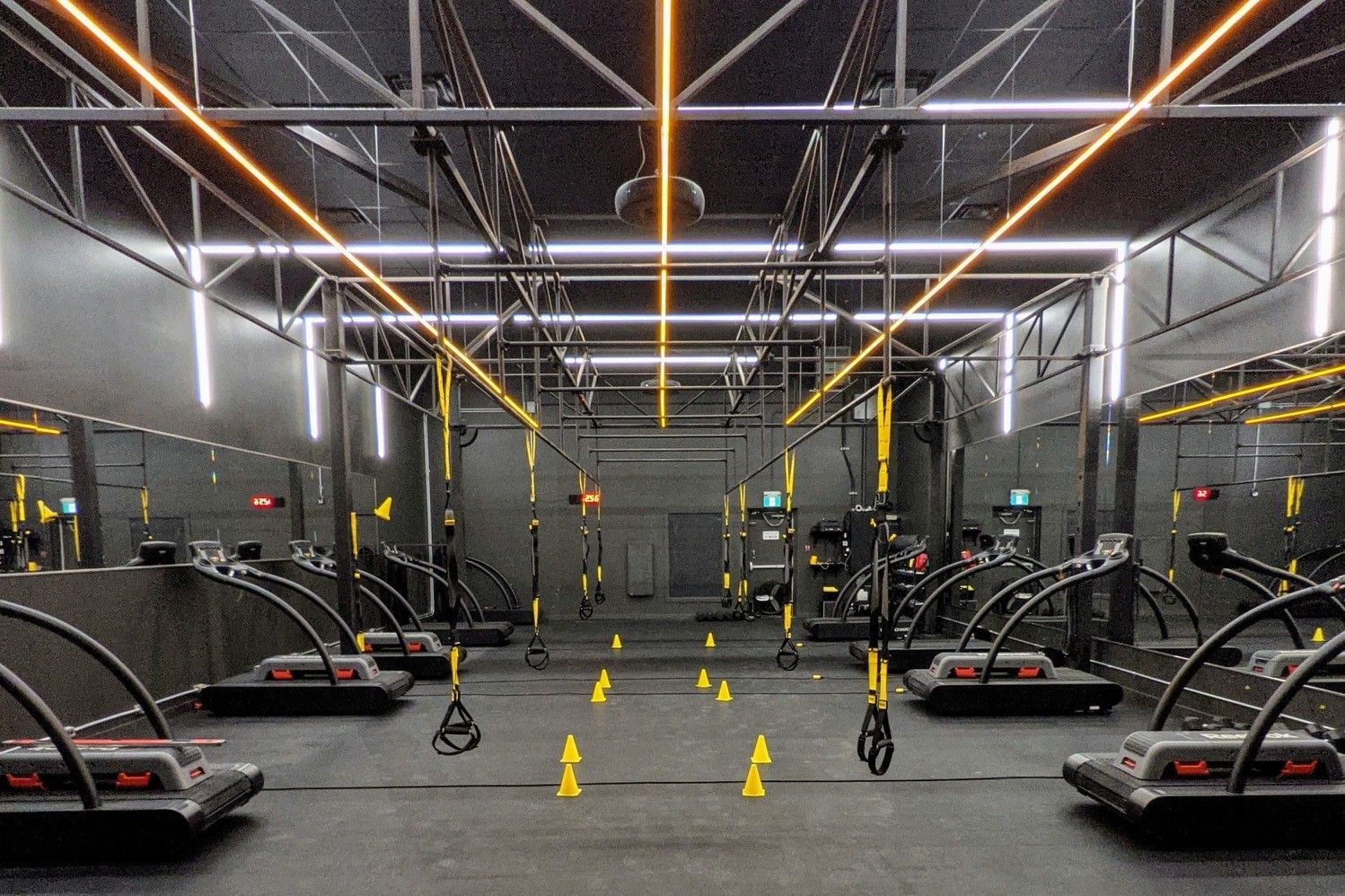 Intérieur de salle de sport avec rangées de machines d'exercice et d'équipements suspendus ; lumières orange et blanches.