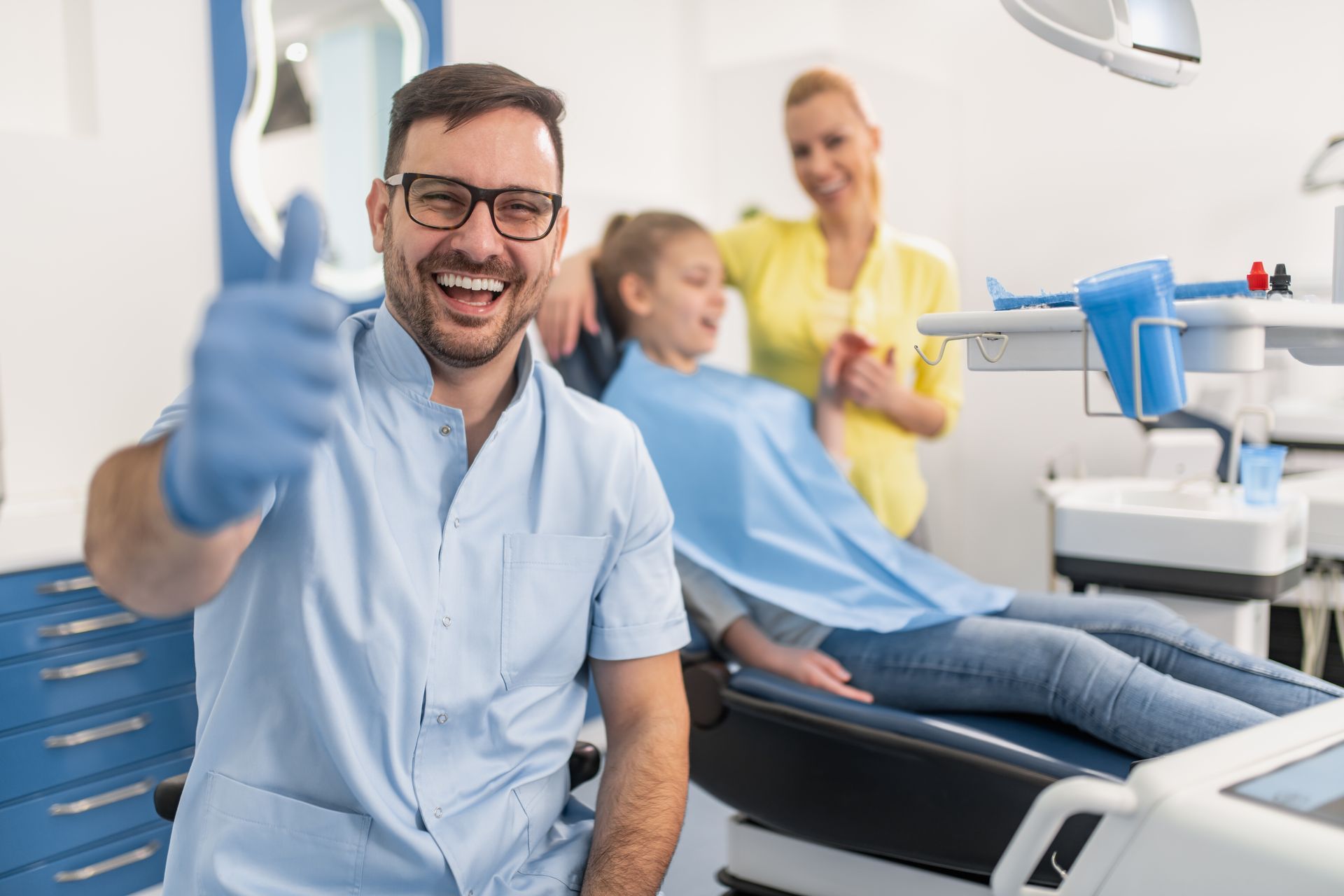 A dentist smiling with his thumb up and a girl sitting in a dentist chair with her mother in the background.