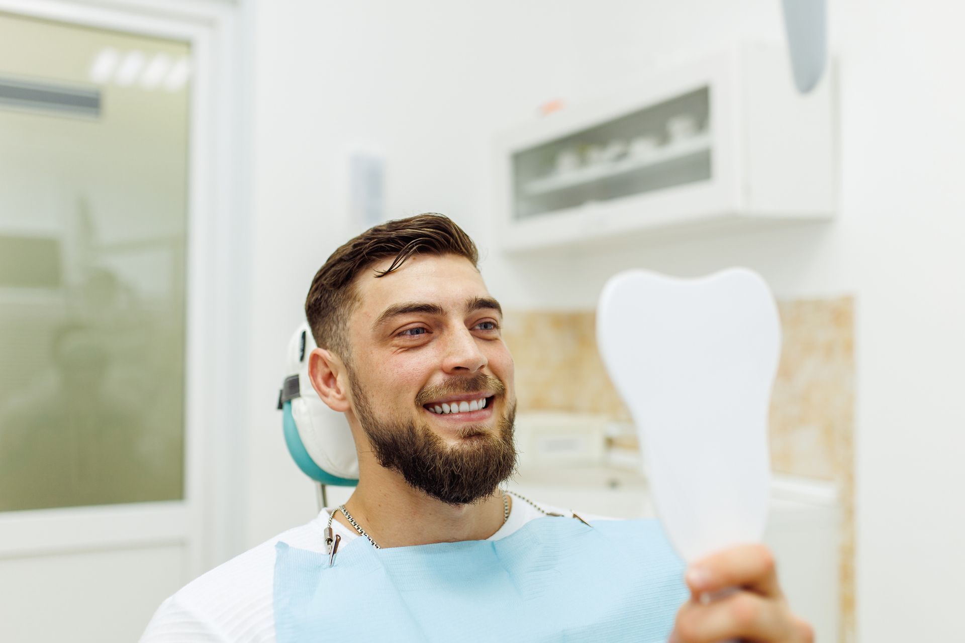 A man is sitting in a dental chair looking at his teeth in a mirror.