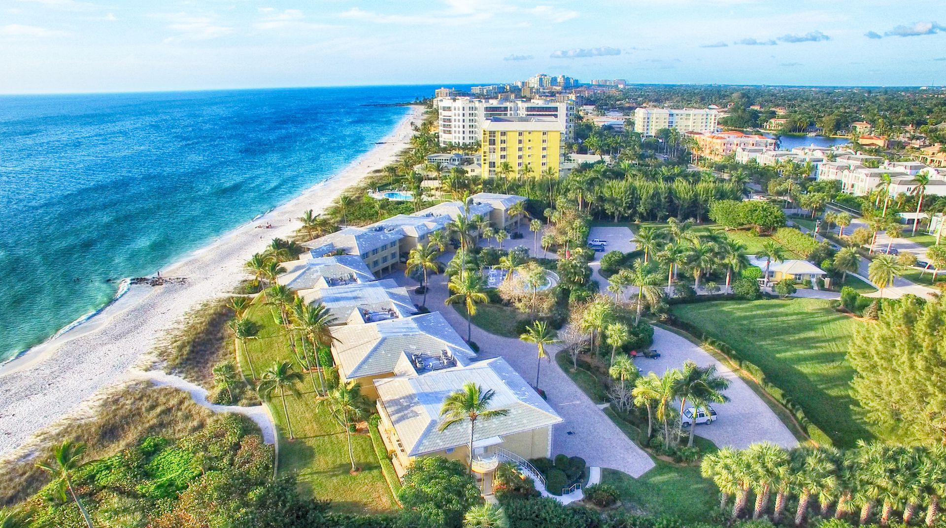An aerial view of a tropical beach resort with multi-story buildings, lush greenery, and the blue ocean along the coast.
