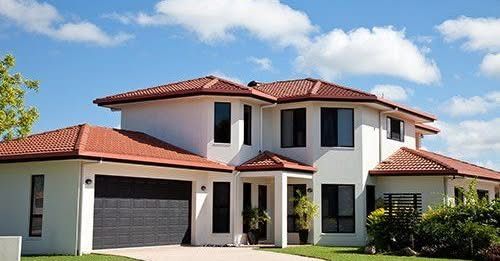 Two-story white house with terracotta roof, black windows, and a dark garage door against a blue sky with clouds.