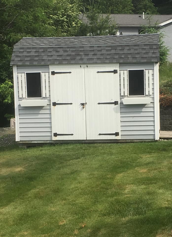 A white shed with a gray roof is in the middle of a lush green yard.