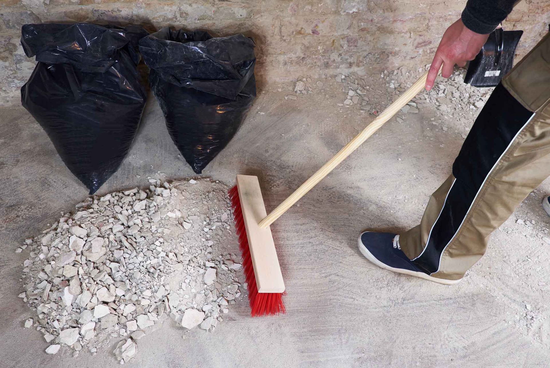 Person sweeping debris into a pile on a concrete floor with two trash bags nearby. Person sweeping debris into a pile on a concrete floor with two trash bags nearby.