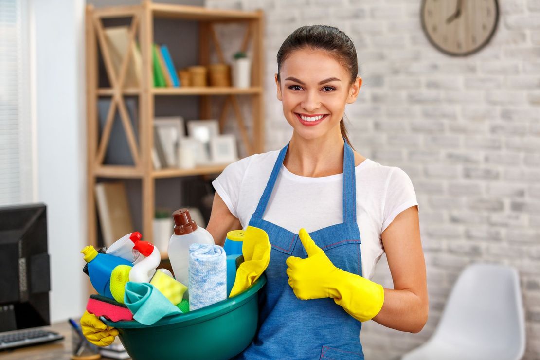 Woman in apron and gloves holding cleaning supplies, giving thumbs up.