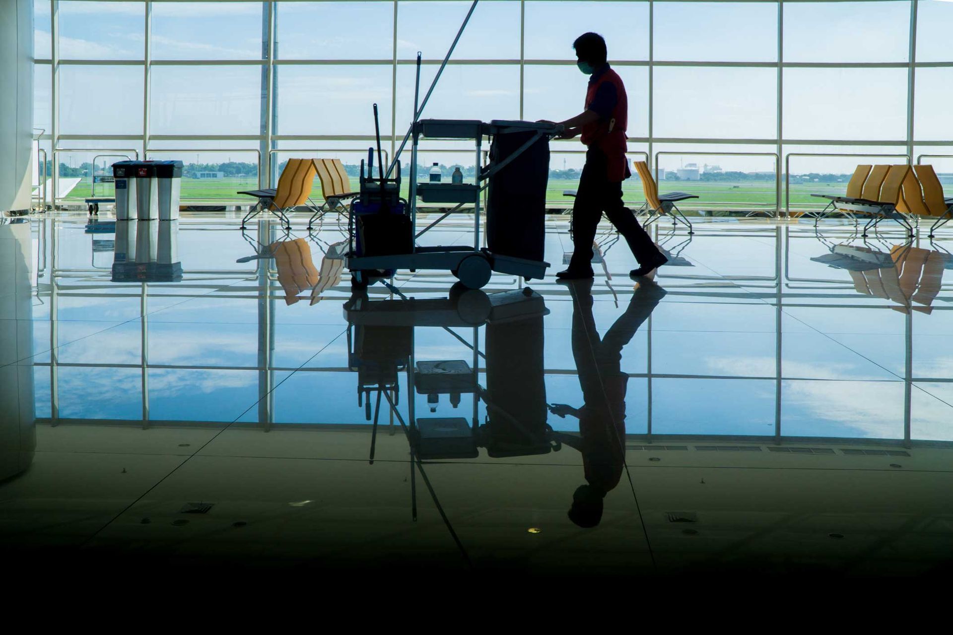 Man in a mask cleaning a large, reflective floor in an airport, pushing a cleaning cart.
