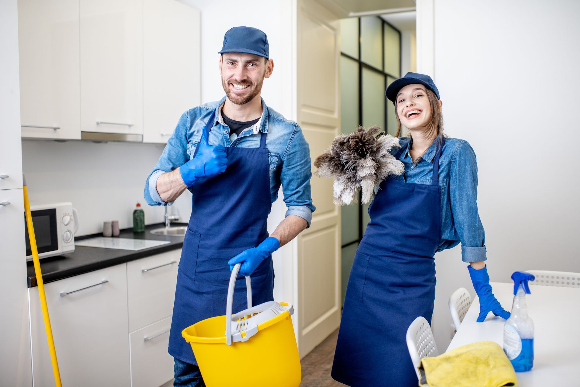 Two cleaners in blue uniforms holding cleaning supplies and a mop in a bright kitchen