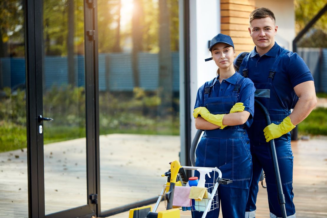Two cleaning service workers, wearing blue overalls, stand by a house entrance with cleaning equipment.