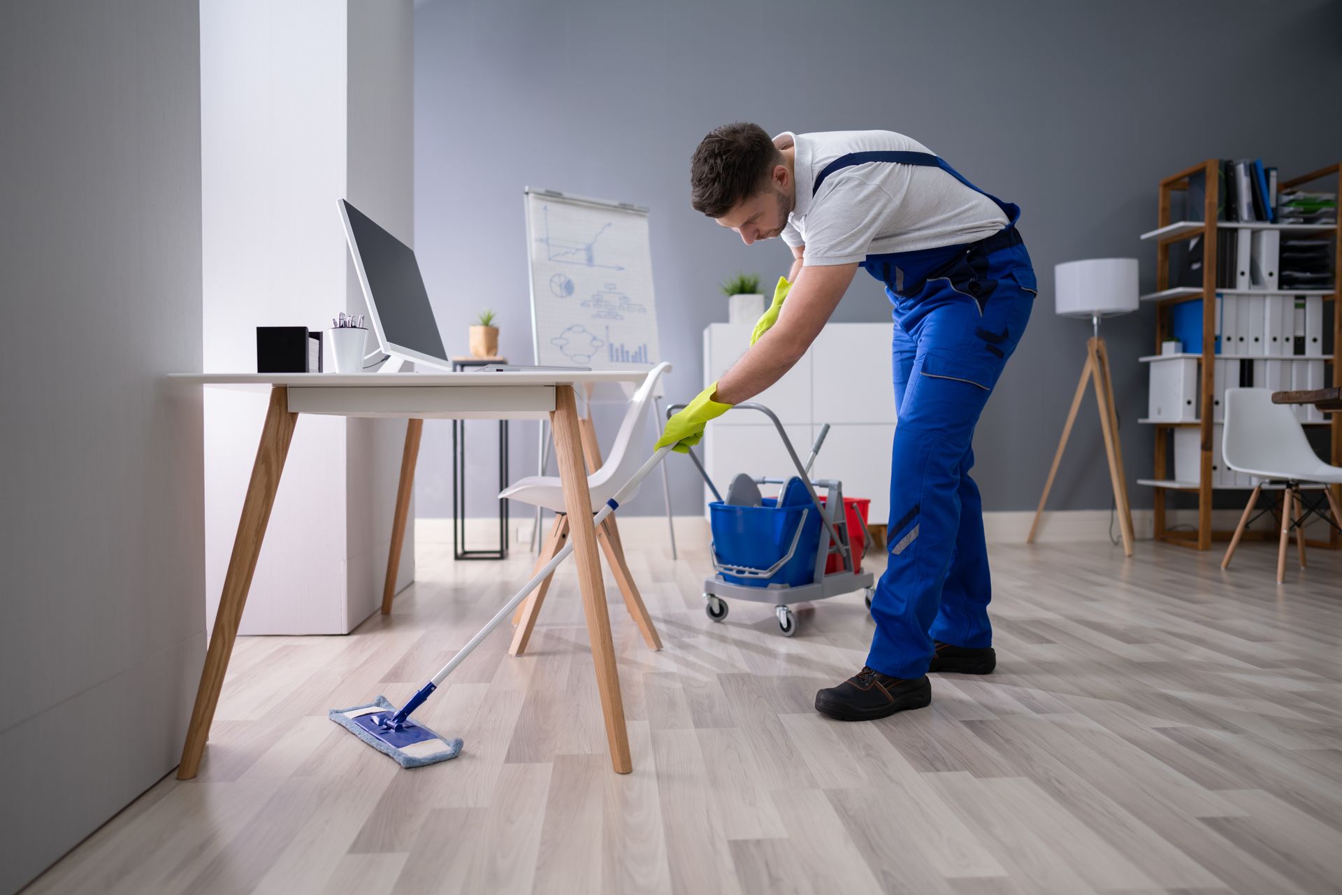 Person mopping a light wood floor in a bright room with a blue bucket and cleaning supplies