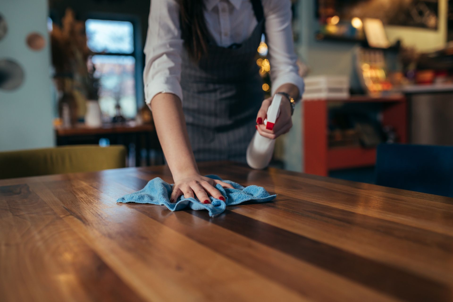 Person wiping a wooden table with a blue cloth and spray bottle in a restaurant.