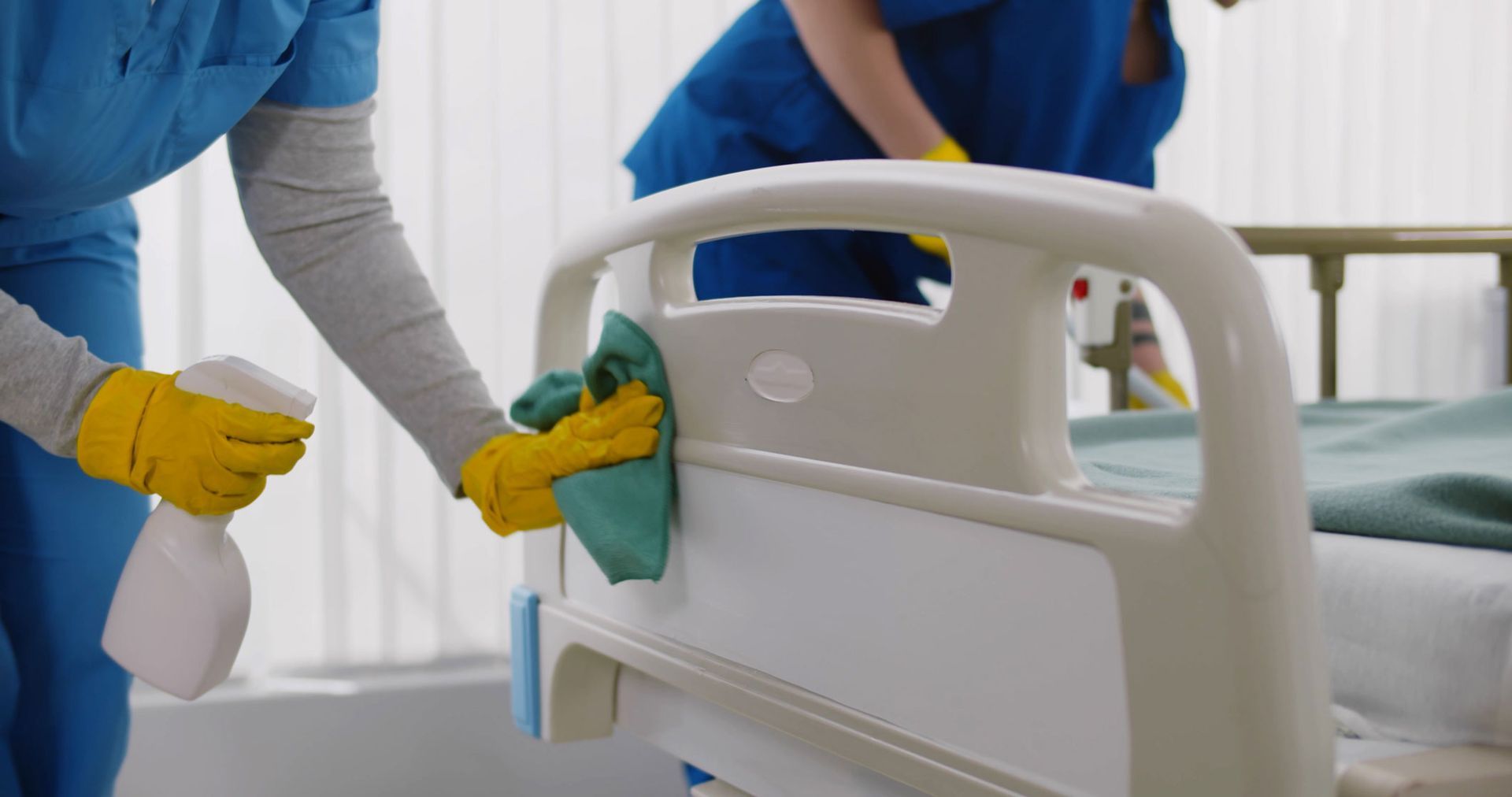 Caretaker wiping a hospital bed rail with a spray bottle and cloth