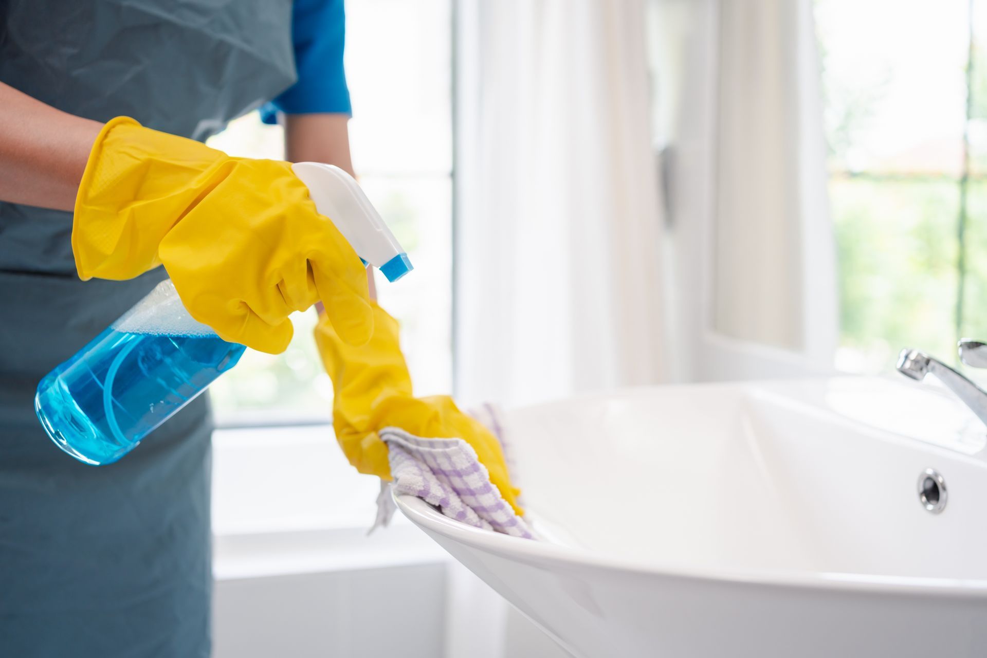 Person in yellow gloves spraying and wiping a white bathroom sink with a spray bottle and cloth. Person in yellow gloves spraying and wiping a white bathroom sink with a spray bottle and cloth.