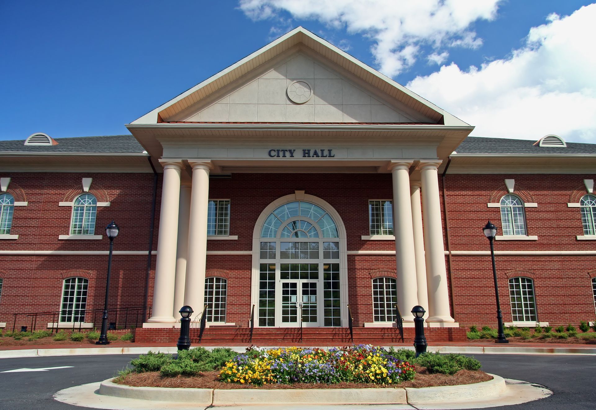 A brick City Hall building with white pillars, a triangular pediment, and a semicircular entrance under a blue sky.