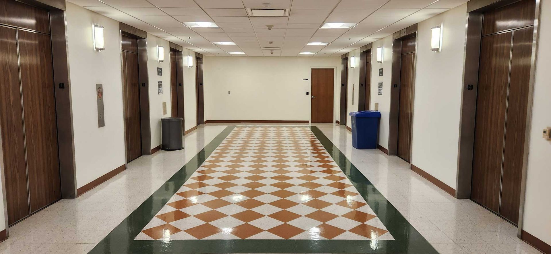 Empty hotel hallway with patterned tile floor, beige walls, wooden doors, and blue trash bins