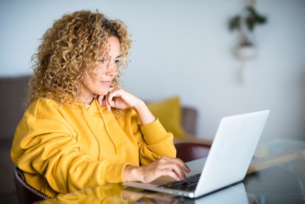 Person in a yellow hoodie using a laptop at a glass table in a bright room