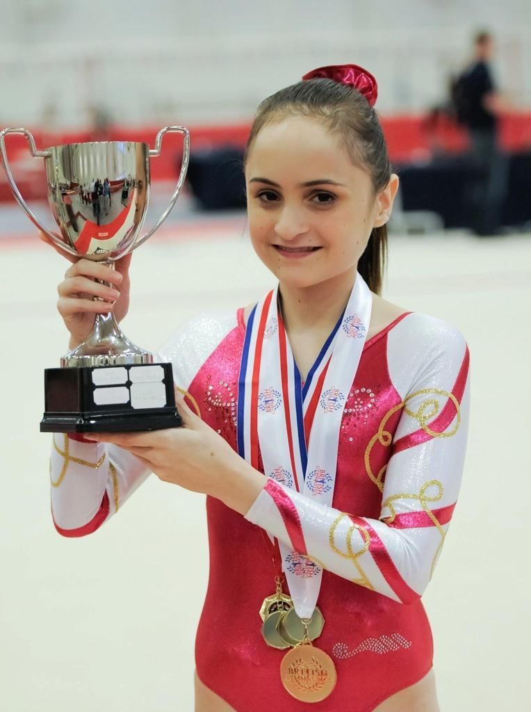 Lauren with brown hair in a ponytail, in a red and white leotard with a silver trophy in her hand and two gold medals around her neck.