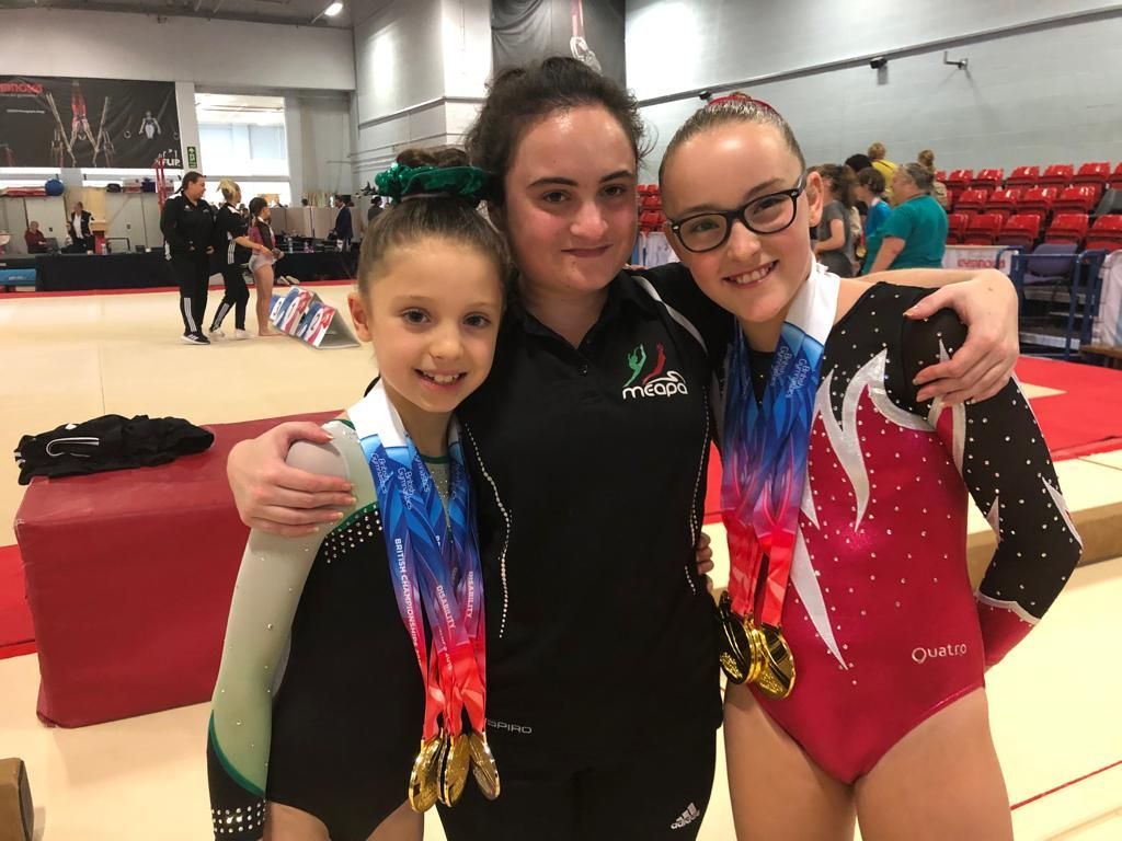 Lauren Lethbridge is in the center wearing a black tracksuit with her dark hair in a ponytail. To her left is Anabelle McAdie in a black and gold leotard with four gold medals around her neck. Sienna Swindon is to her right and is wearing a black, red, and white leotard and has three gold medals around her neck. 
