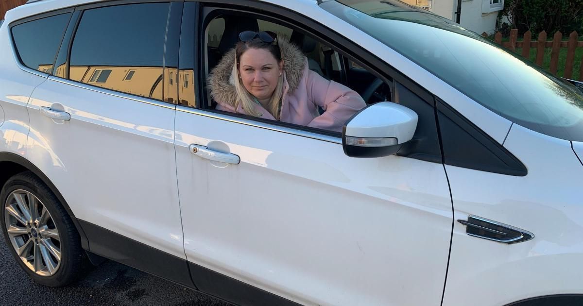 Victoria Jayne with blonde hair and wearing a pink coat, leans out the window of the drivers seat of a white car. 
