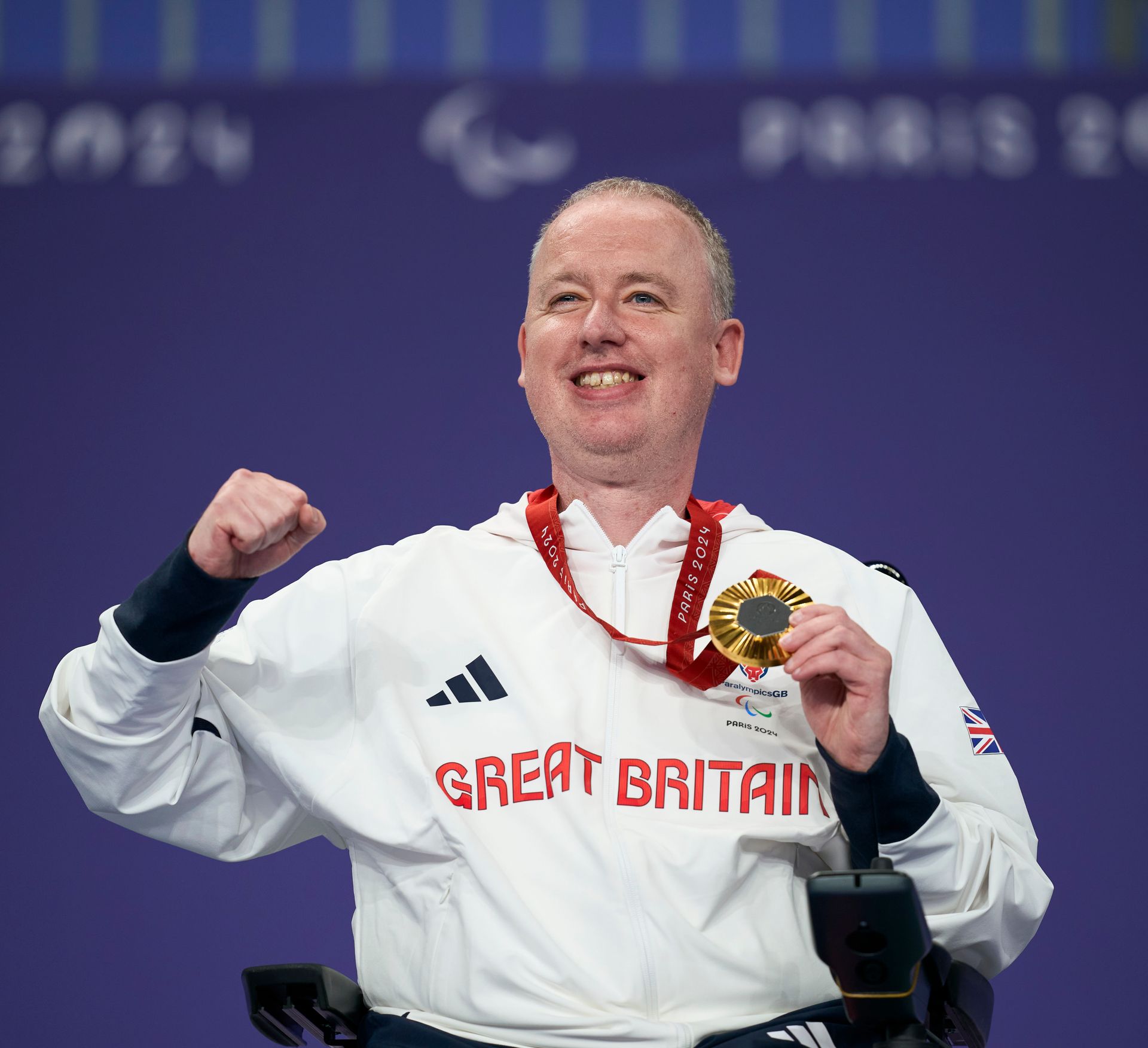 Stephen has grey hair and wears a beaming smile. He has a white jacket on with Great Britain in red writing across the chest. A gold medal on a red ribbon hangs around his neck which he holds in his right hand. His left hand is punching the air. 