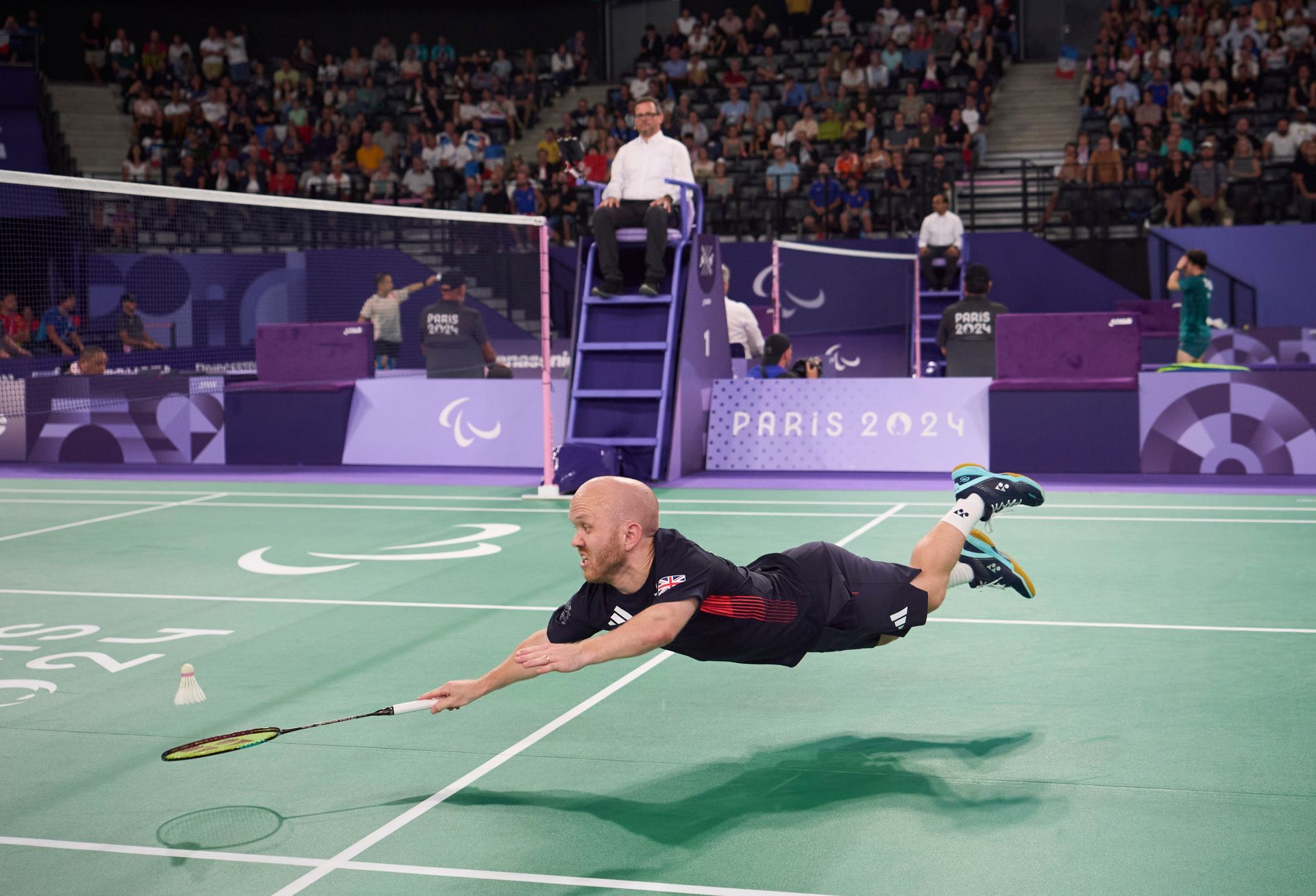 Krysten who is bold and wearing a black t-shirt, black short and blue shoes and white socks. He is flying through the air in an horizontal position with a racket in his left hand. He is on a badminton cour and a purple side of the arena are in the background with Paris 2024 written on it. 