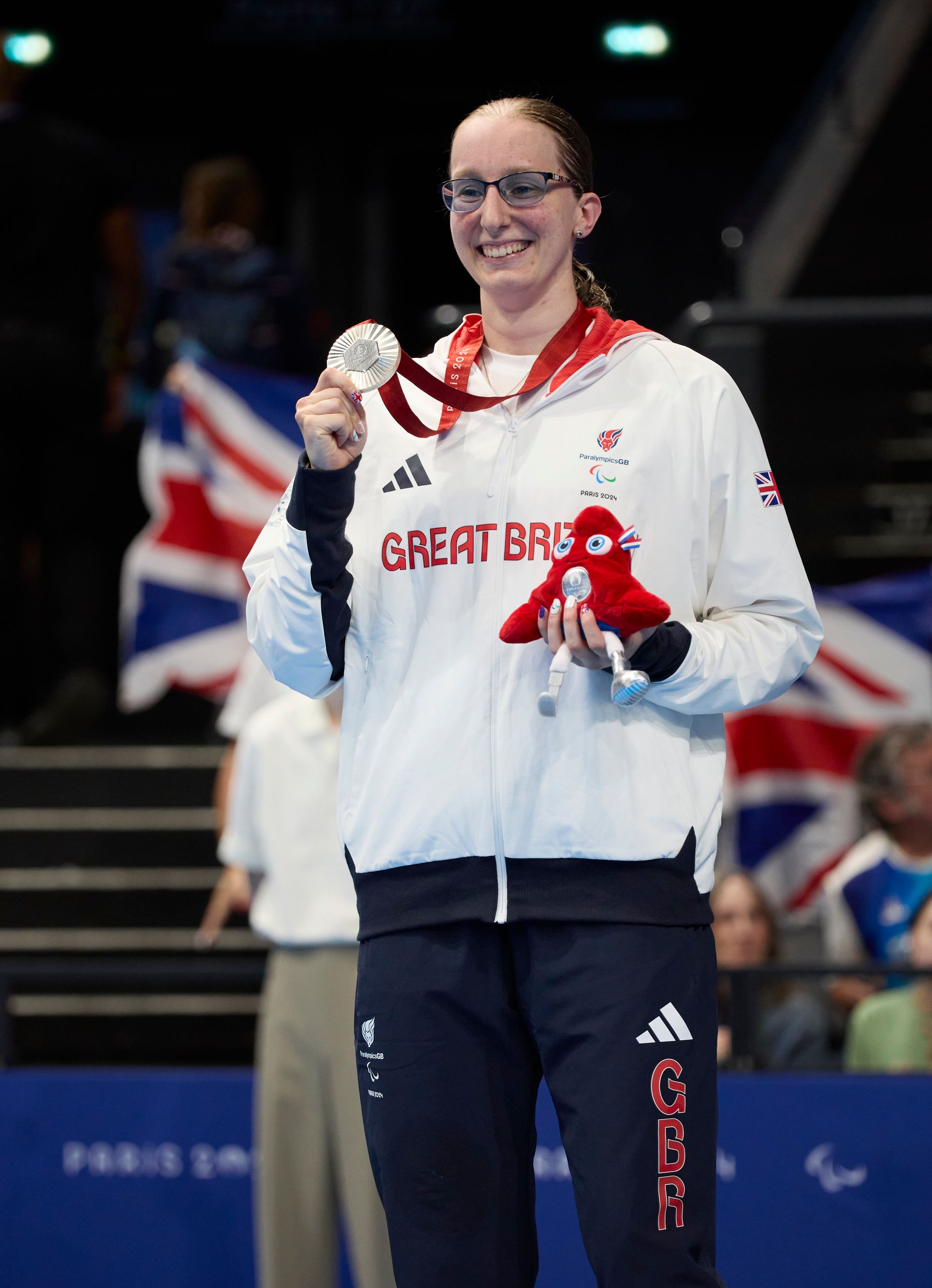 callie-ann has brown hair and wears glasses. she has a white jacket on with Great Britain in red writing across the chest. She has blue tracksuit bottoms on with GBR up the right leg and the Adidas logo. She is holding a silver medal in her right hand on a red ribbon around her neck. In her left hand she has a red cuddly toy. 