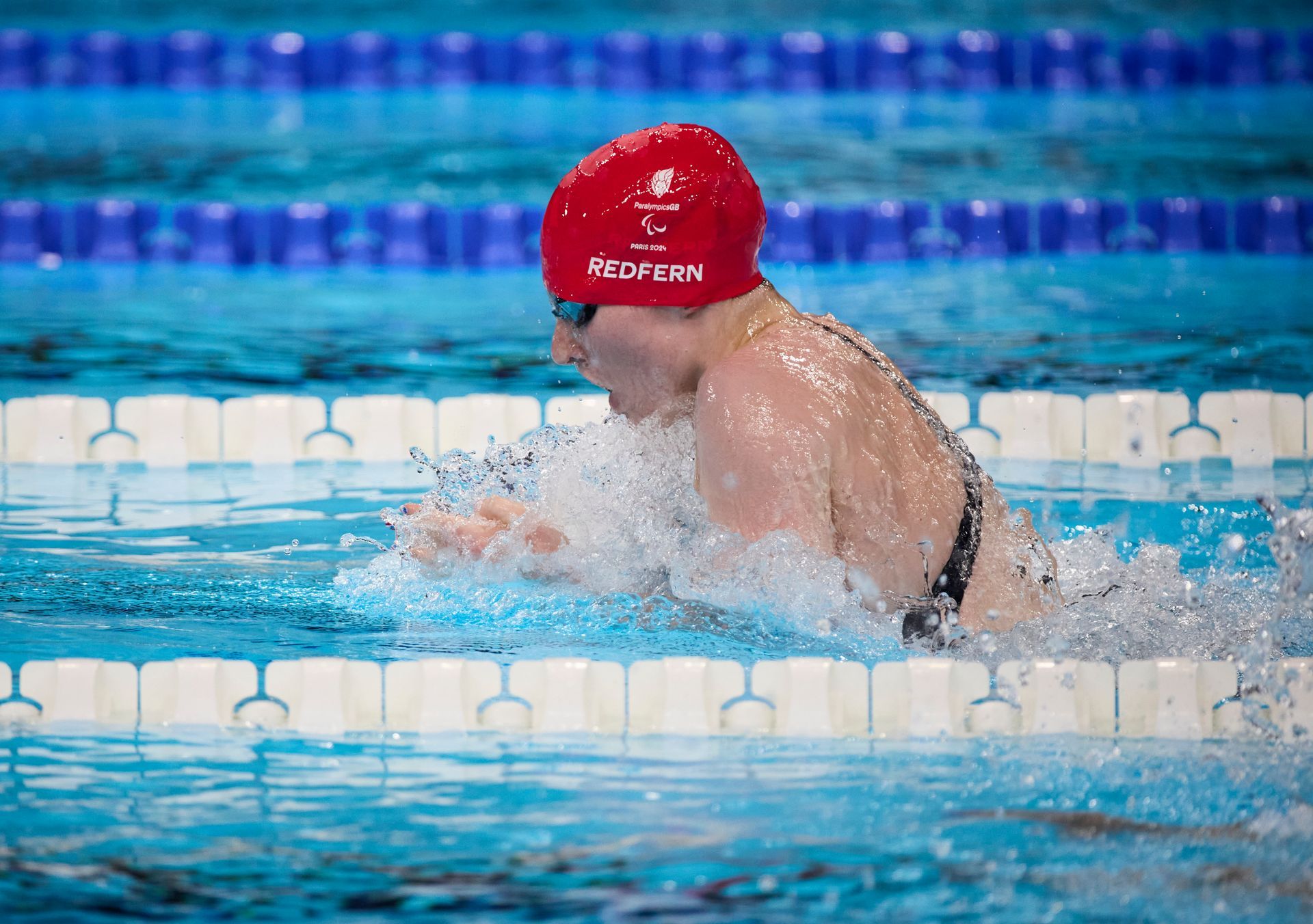 Rebecca has a red swimming hat on, with the words REDFERN on with the ParalympicsGB name and motif. She is wearing swimming goggles and a black swimming costume. She is swimming in the water with water splashing up in he face. In the distance, blue and white ropes separate the lanes. 