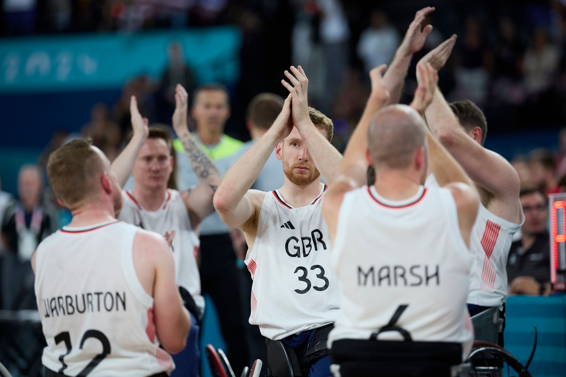 The Mens GB team sat in their wheelchairs in white vests. the words Warburton 12 Marsh 6 and GBR 33 can be seen on three of the shirts, the others are in the distance. 