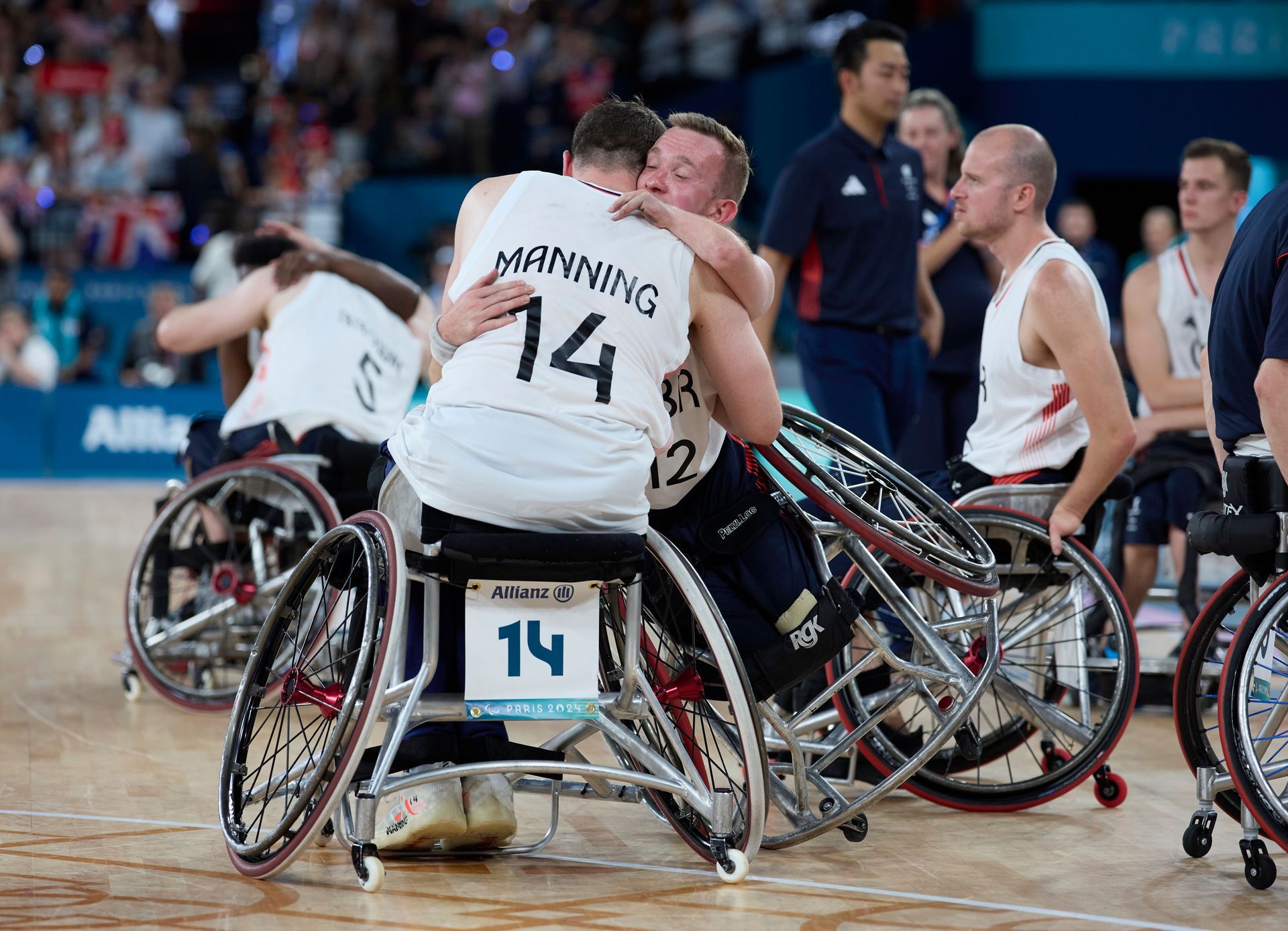 Manning with dark hair and Warburton with blondish in their wheelchairs hugging each other. Both are in white vests Manning who has his back to the picture has the words Manning on his top and the Number 12. Warburton who is towards the picture you can see the number 12 on his shirt. other players are in the background in white vests and members of the public