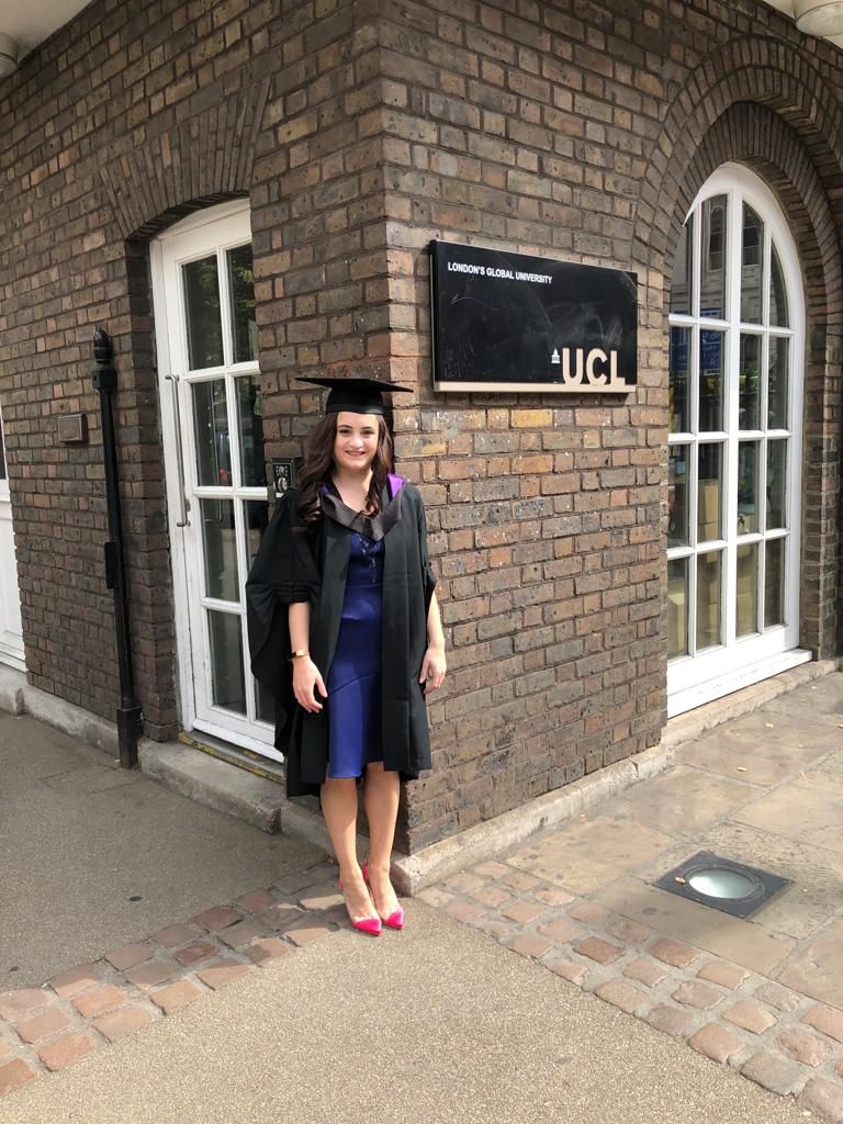 Lauren Lethbridge in front of a brick wall and a sign which read UCL. She is wearing a purple dress black greaduation gown and mortarboard hat and pink shoes. 