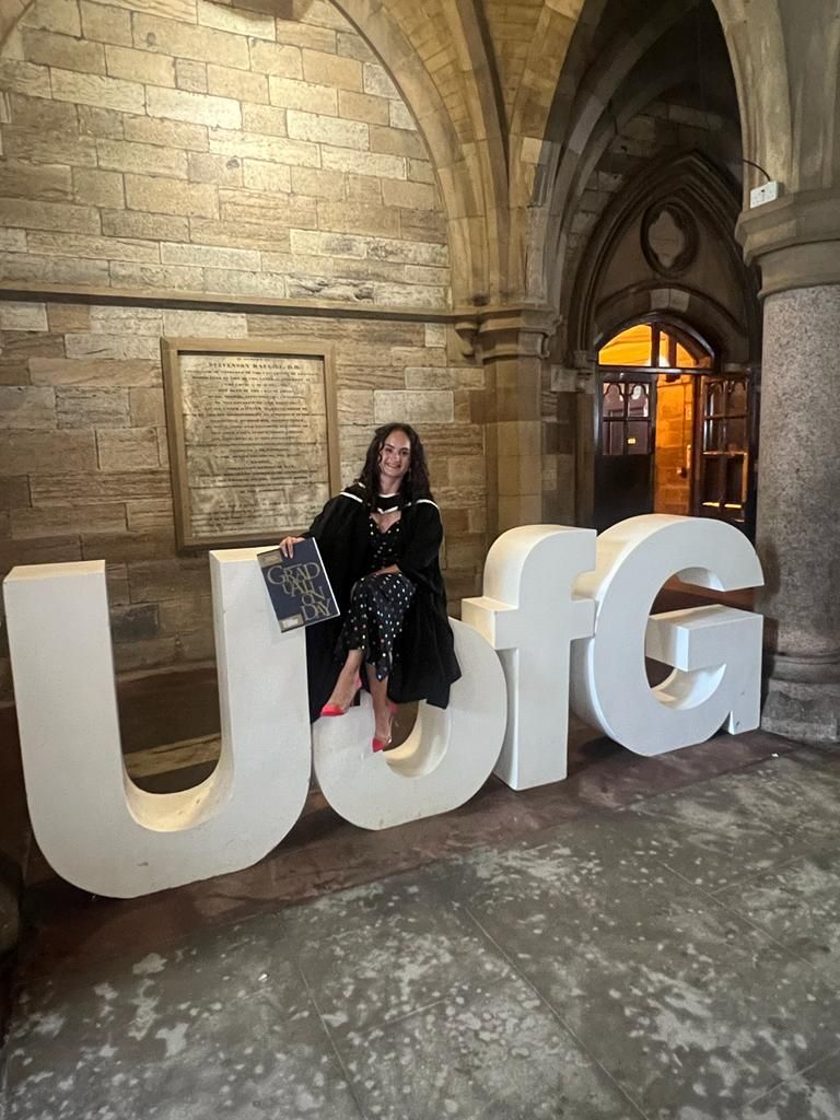 Lauren Lethbridge is weraing a spotted dress with her black graduation gown over the top and black and white hood with pink shoot. She is holding her graduation programmer in her right hand and is sitting on the UofG letters which are big and white. 
