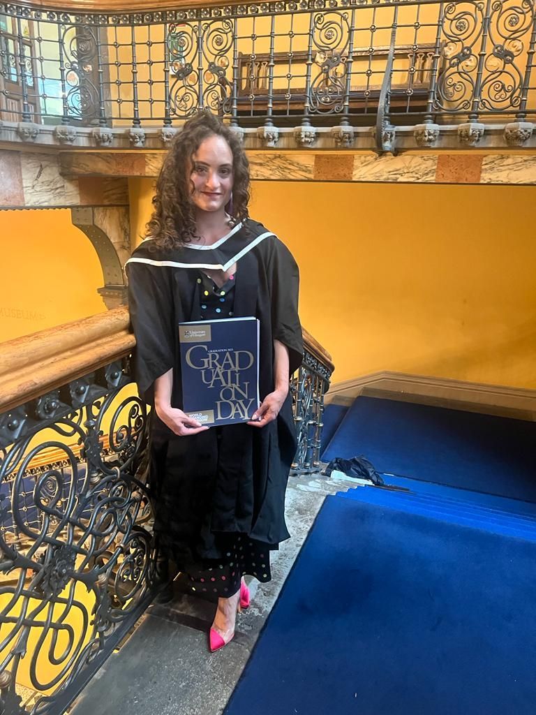 Lauren Lethbridge in a black graducation gown with black and white striped hood. SHe is wearing a black dress and pink shoes. SHe is standing on a landing which is painted yellow and has blue carpet with a black iron staircase. 