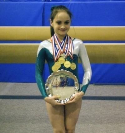 Lauren wearing a green and white leotard with five gold medals around her neck and holding an award