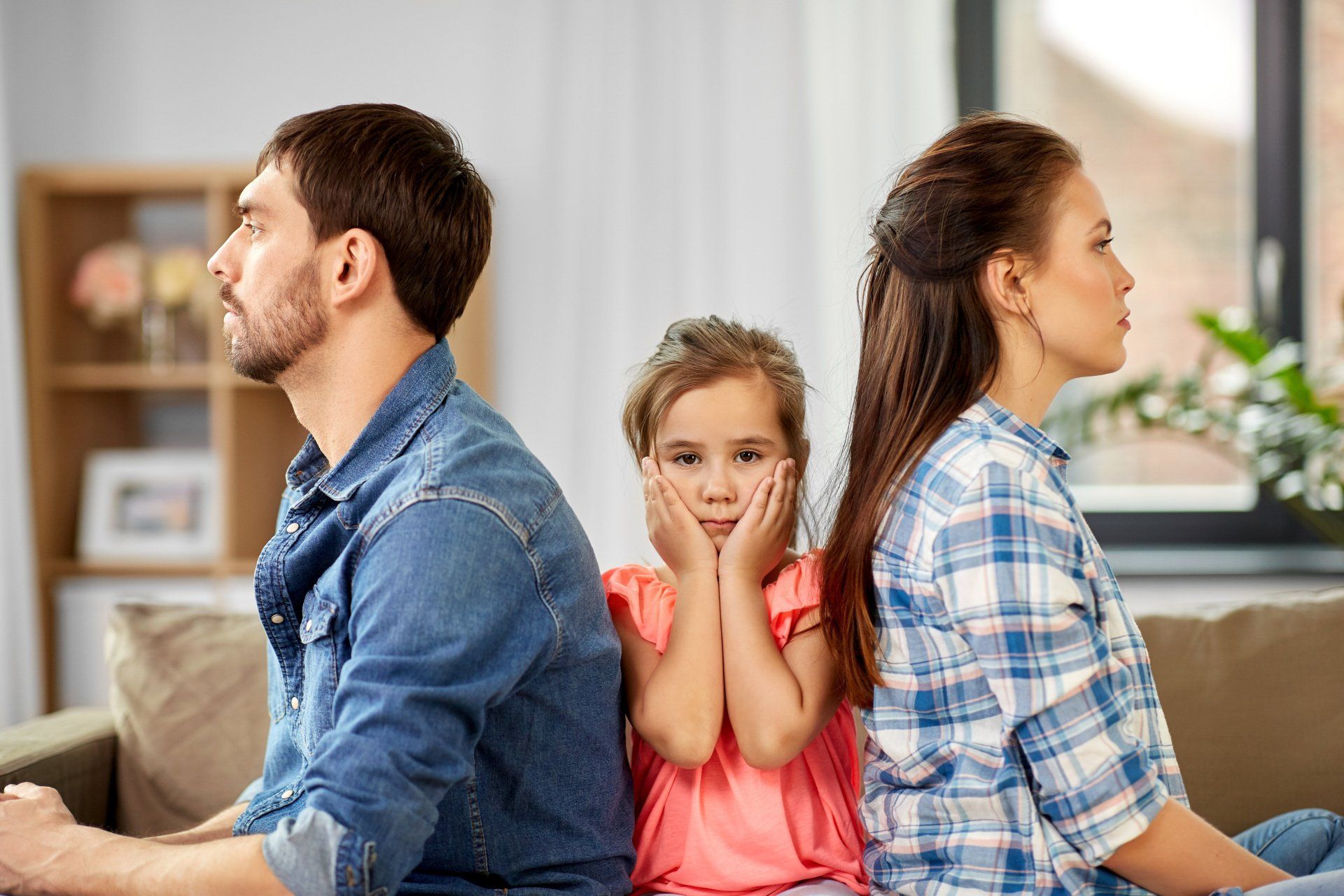 Man look right dark hair in a denim shirt. Women looking to the right dark hair checked shirt. A little girl sits in the middle looking straight on with her head in hands. SHe has a peach top on with mousey hair.