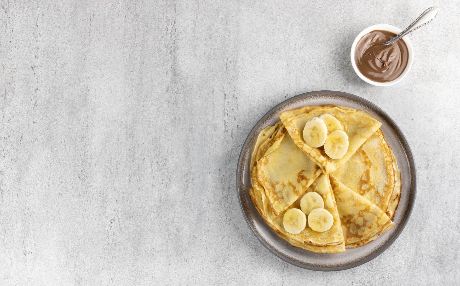 pancake on a round dish topped with sliced bananas. in a seperate bowel is melted chocolate with a silver spoon stuck out,
