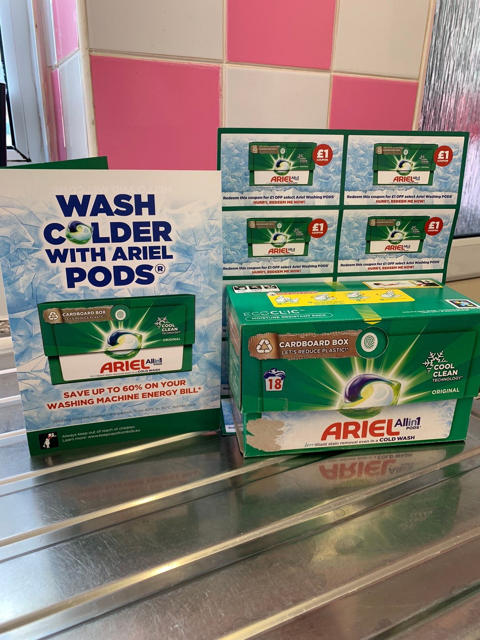Box of ariel washing tablets in a cardboard box  with a leaflet stood up next to it say wash colder with ariel pods save upto 60% on your washing machine energy bill. Behind the ariel box is some cupons for £1 off your next box. All of these products are stood on a silver draining board with pink and white tiles in the background.