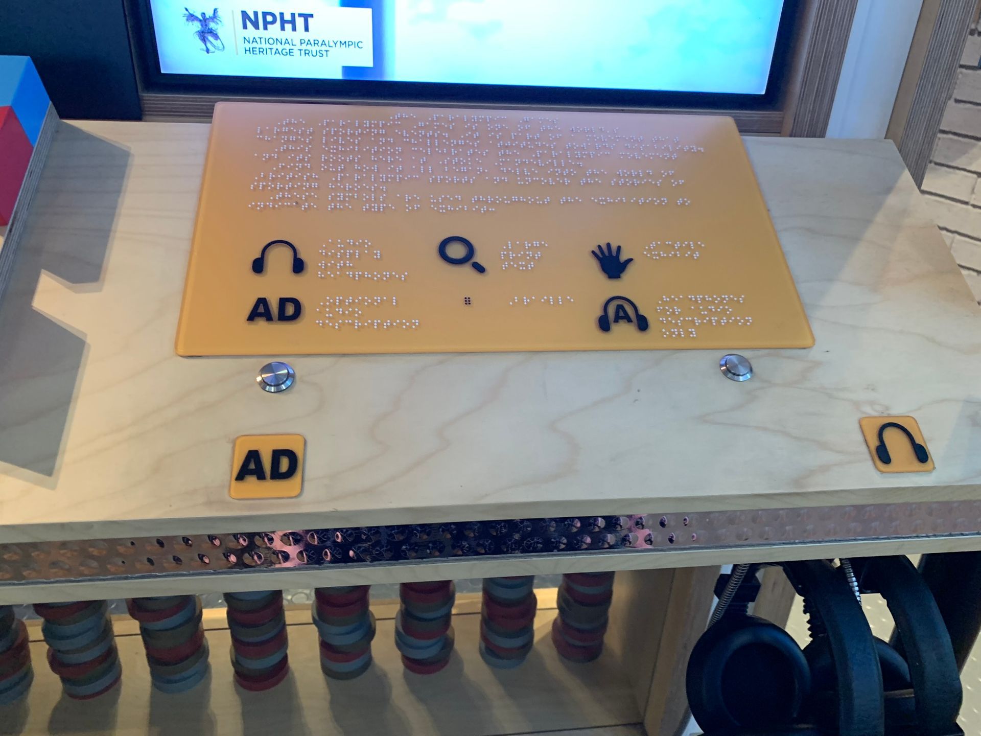 yellow plaque with white braille. The letter AD and a headphone symbol, a magnifying glass symbol, a hand symbol, and an A in a pair of headphones with a screen in the background. 