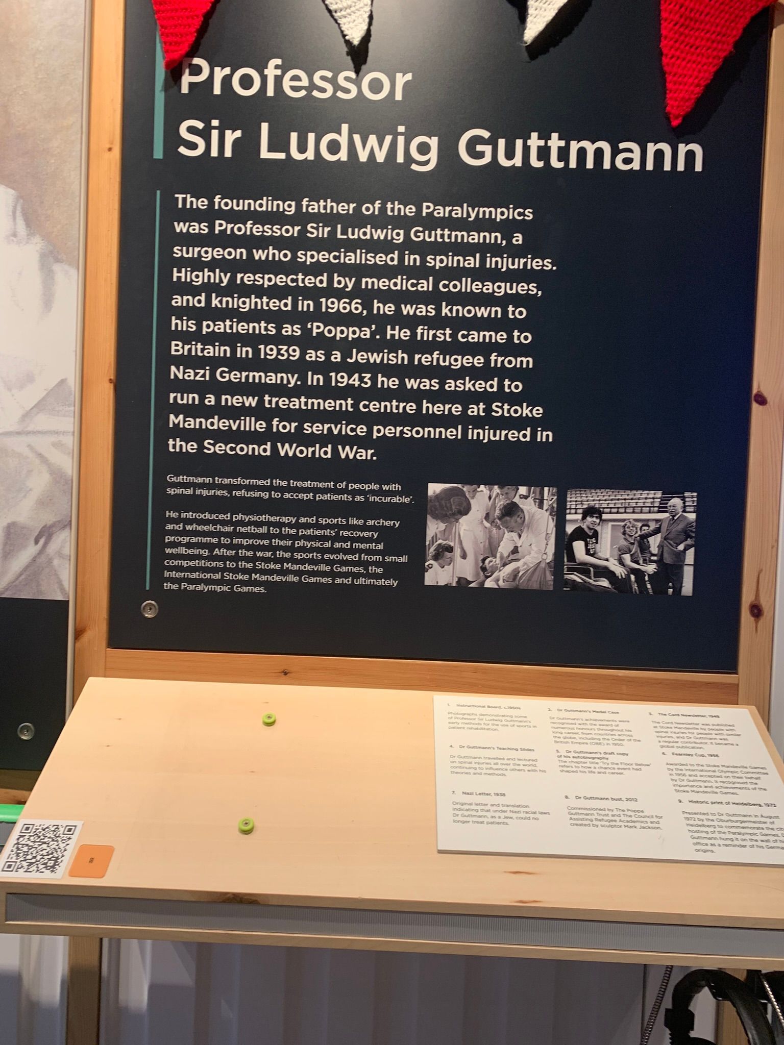 Black poster with writing about the history of Ludwig Guttman and pictures of him in his early years on the side. At the front is a board for people to learn on and write along with a QR to scan to listen to an audio version of the text. 