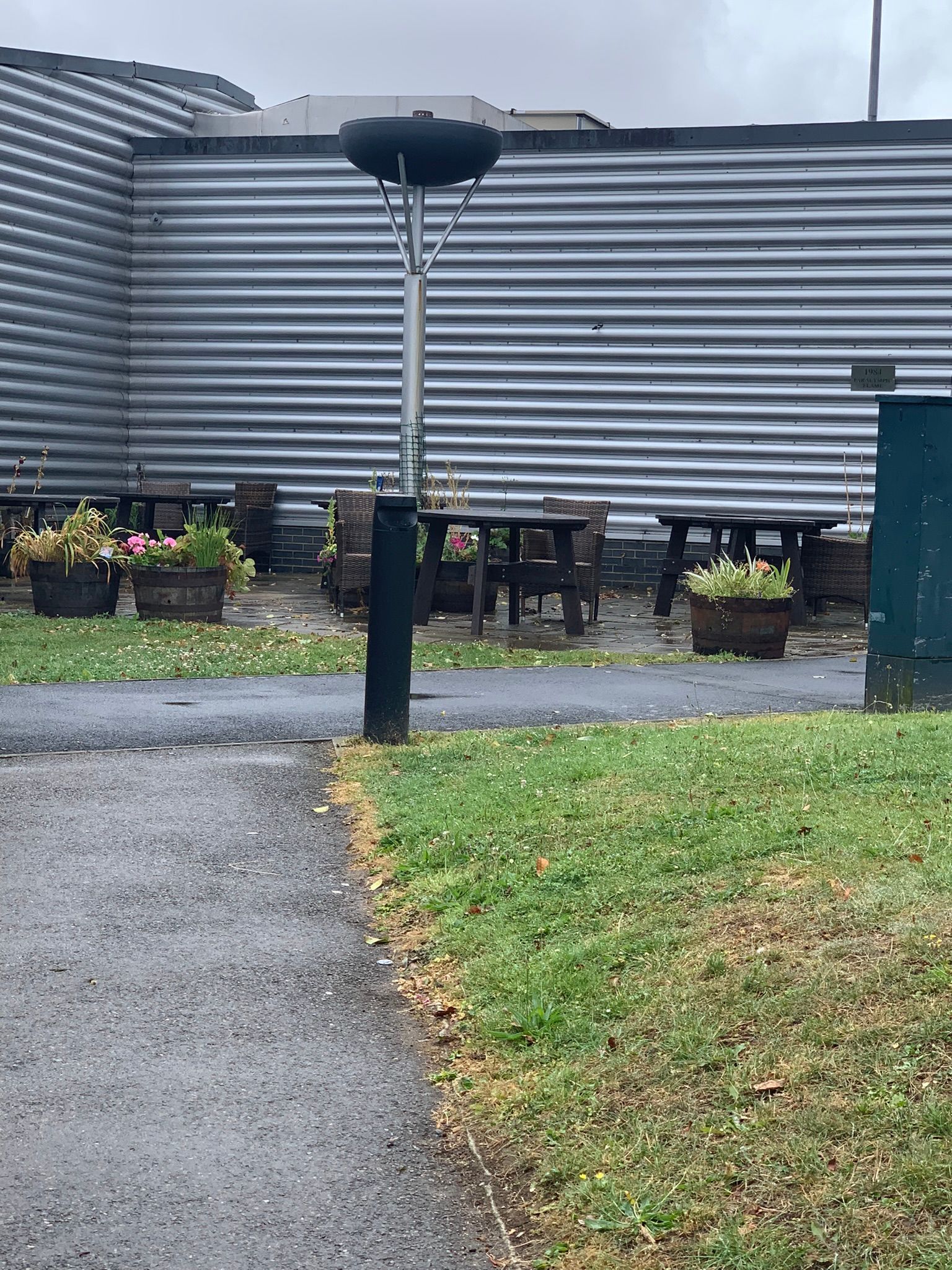 Black cauldron on a metal pole, situated on the edge of a pathway and green area with a sports hall in the background.