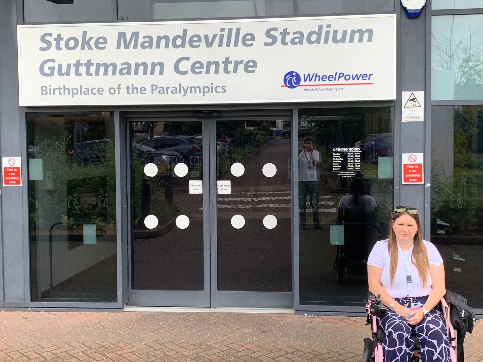 Victoria-Jayne is sitting in her pink wheelchair, with blue and white patterned trousers and a white t-shirt. Long dark hair with sunglasses on her head. Double doors in the background with a sign over the top saying Stoke Mandeville Stadium, Guttman Centre. Birthplace of the Paralympics. 