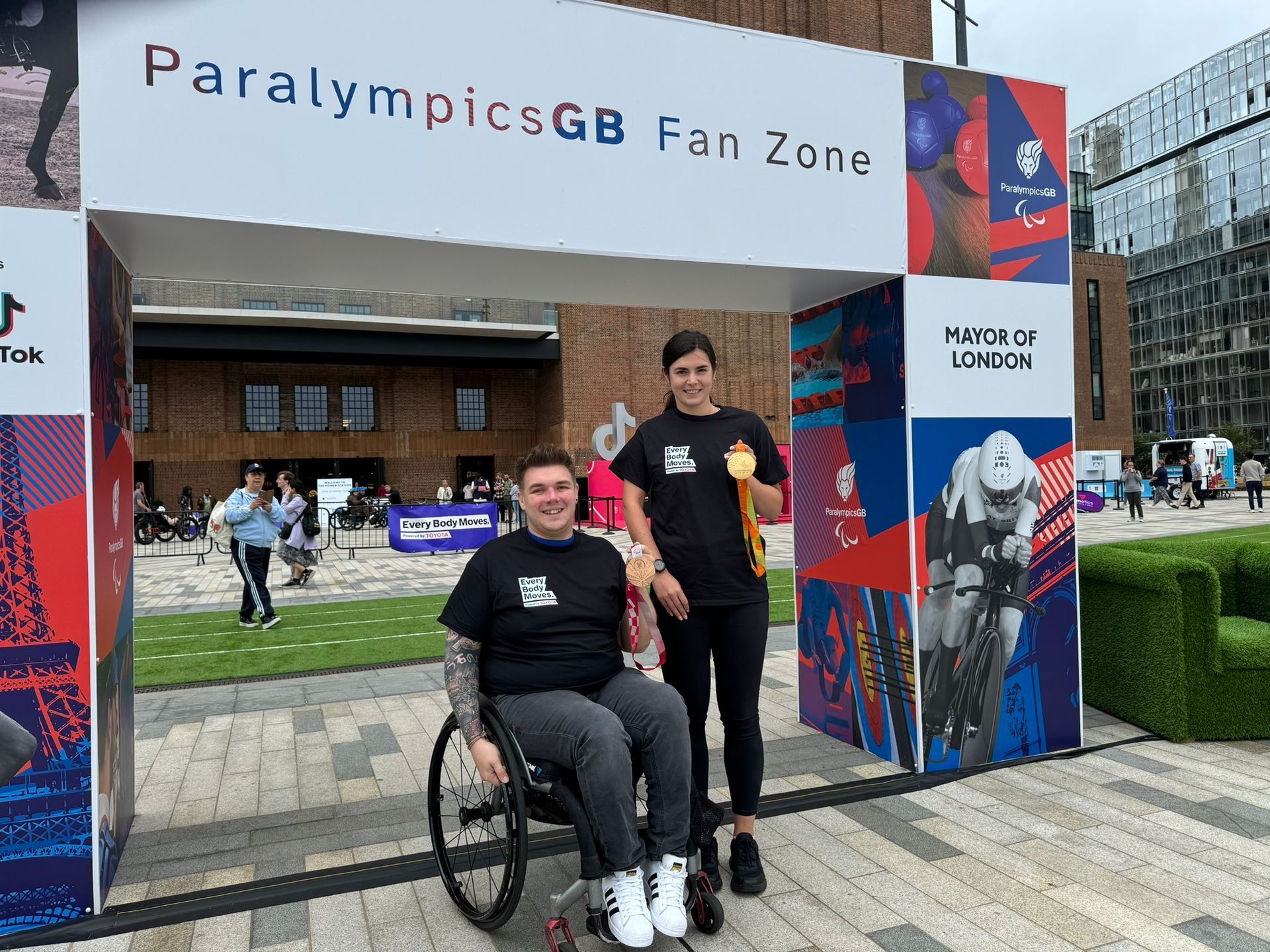 Jack has dark hair and is wearing a black t-shirt with the everybody moves logo and black leggings. He is sat in his wheelchair and holding his gold medal in his left hand. Grace is stood to his left in a black t-shirt with the everybody moves logo and black leggings she is holding her gold medal in her left hand. They are standing under an arch that says ParalympicsGB Fan Zone. In the background is a grass racing track and a building. 