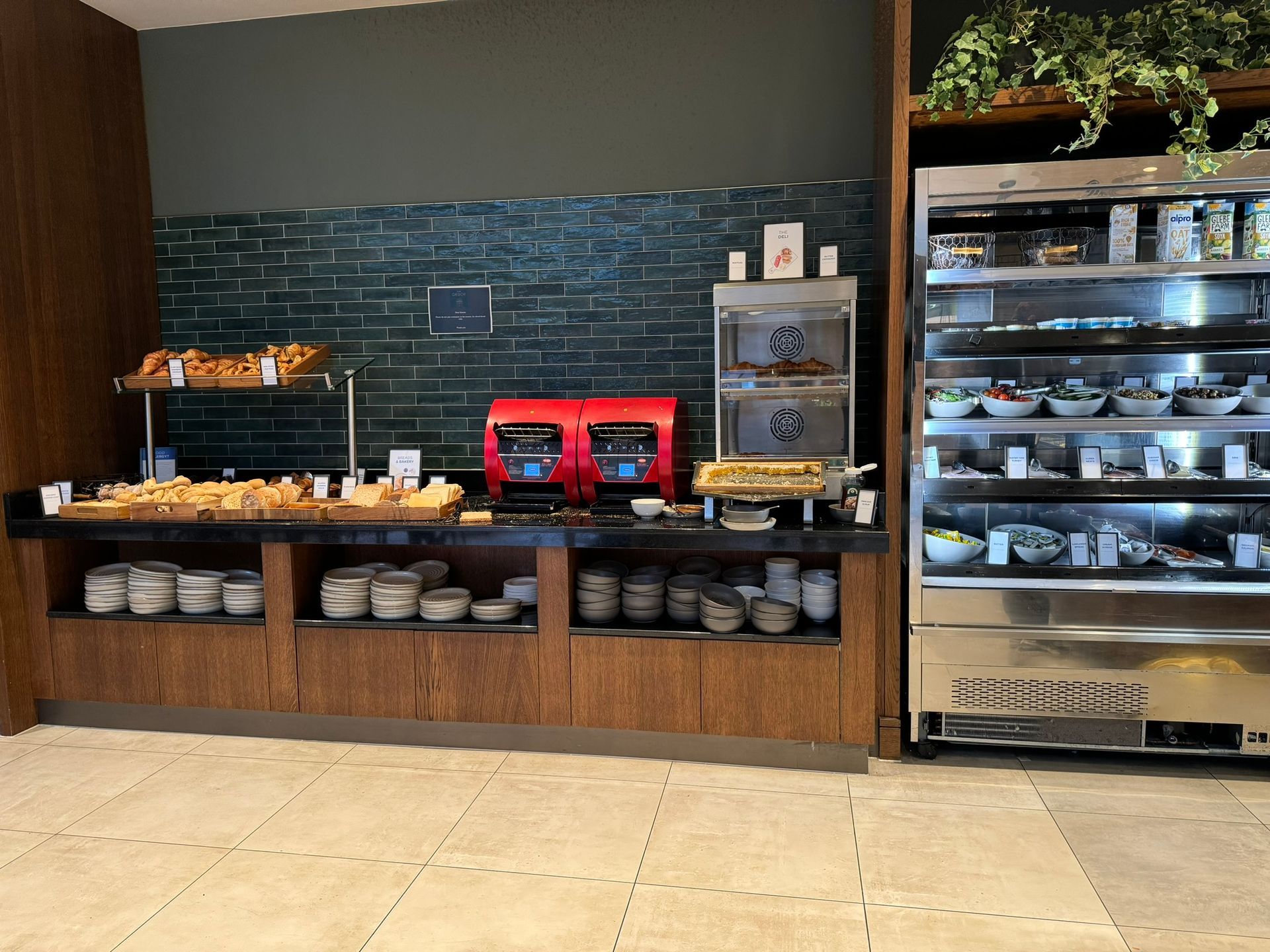 A fridge holding a selection of yoghurts, fresh fruits, ham, cheese and fish. To the left a waffle make and toasters. Underneath the unit sits a selection of bowls and plates. 