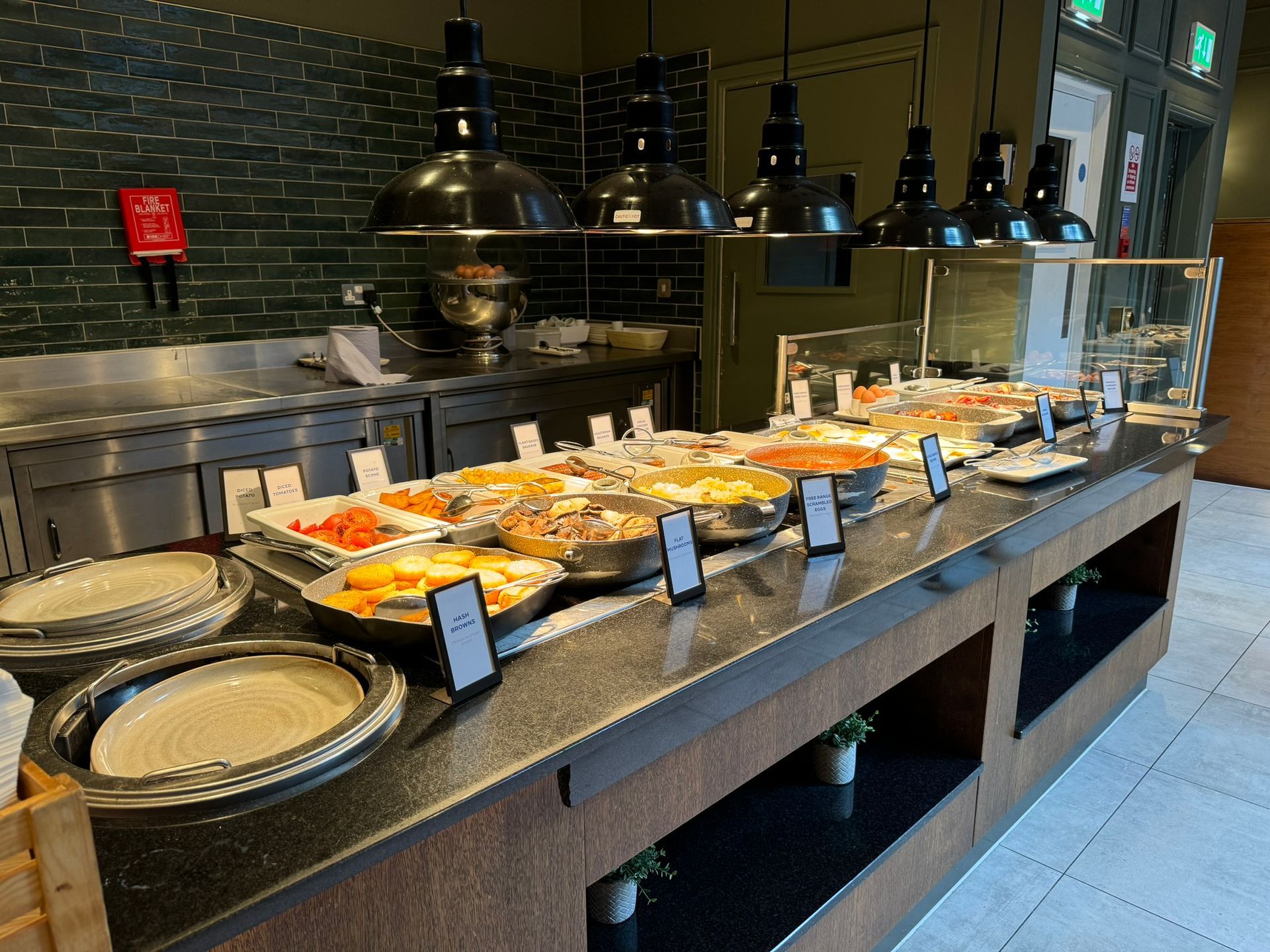 A black brick wall sits to the background with a steel counter in front. In front of that, a hot counter with 6 black downlighters that cover a variety of different hot foods including, mushrooms, tomatoes, bacon, sausages, baked beans, and eggs. to the left of the food is a stack of plates. Pass the food is a white door and next to it is a grey door with a fire exit sign over the top. 