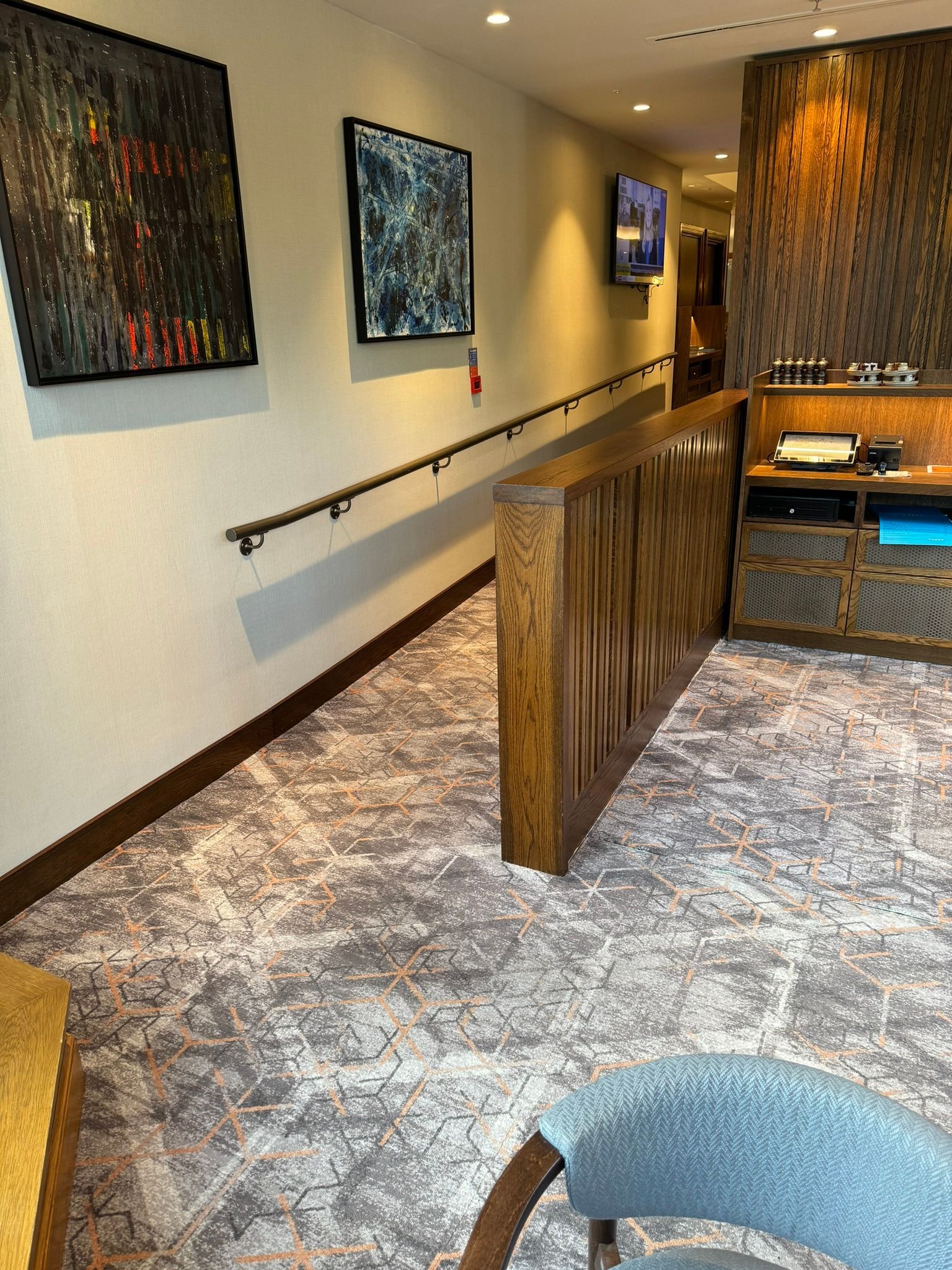 ramp with a handrail on the left and a half-sized oak wall on the right, it is a carpeted area with seas. Two Abstract pictures hang on the wall. 