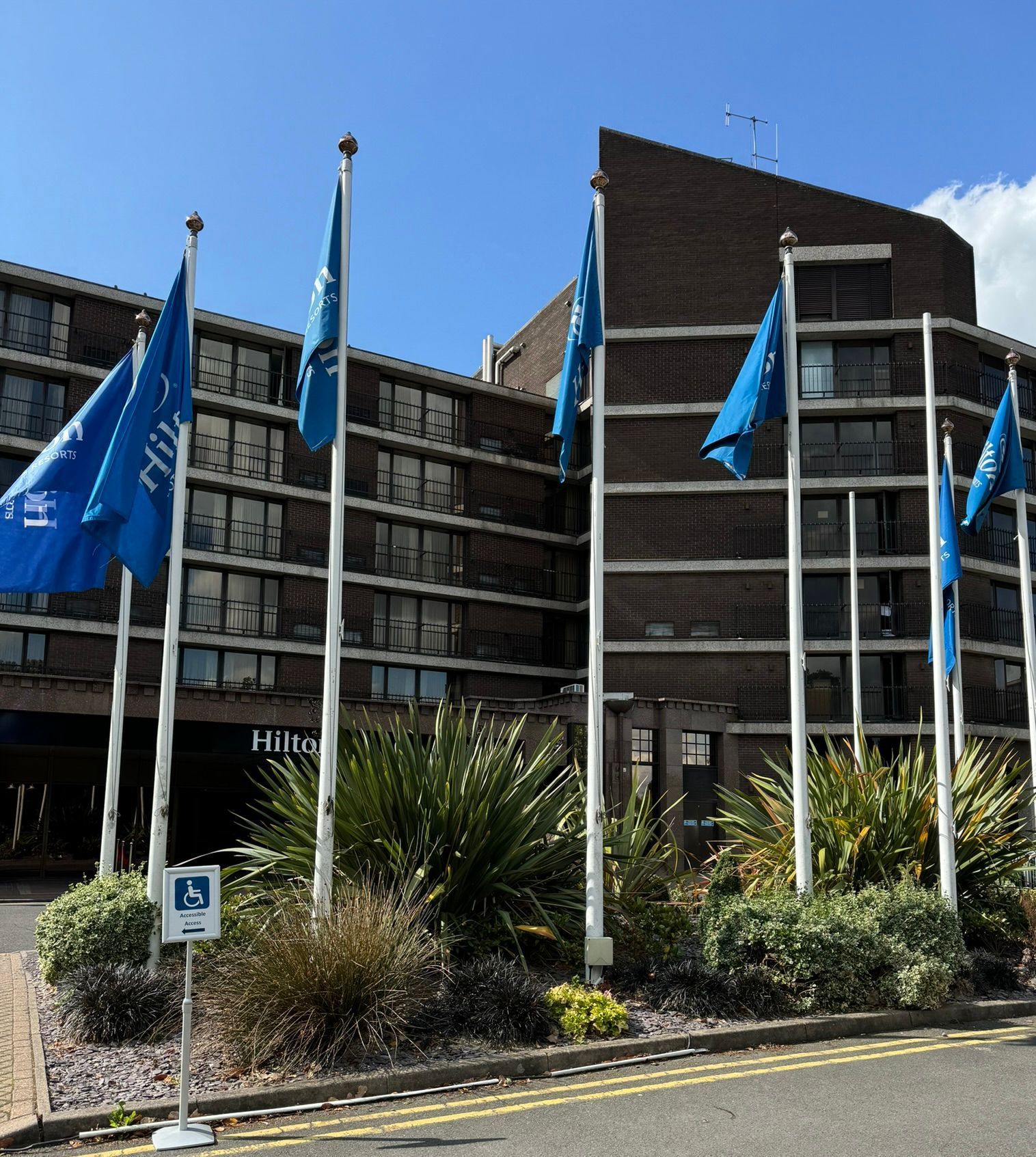 5-story brown brick hotel in the background with white Hilton signage. Plants line a pathway. big and small. 7 flag poles stand in front with blue flags and the Hilton logo and Hilton name adorn them. A disabled sign saying disabled entrance this way sits to the left of the picture.  